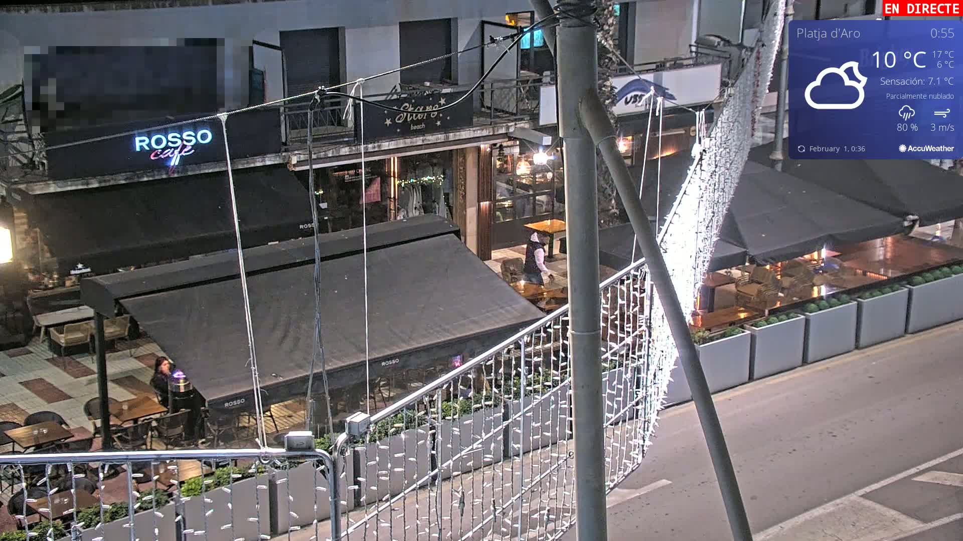 A nighttime, partially cloudy street scene shows several outdoor cafes with dark awnings and string lights, some with people seated at tables.
