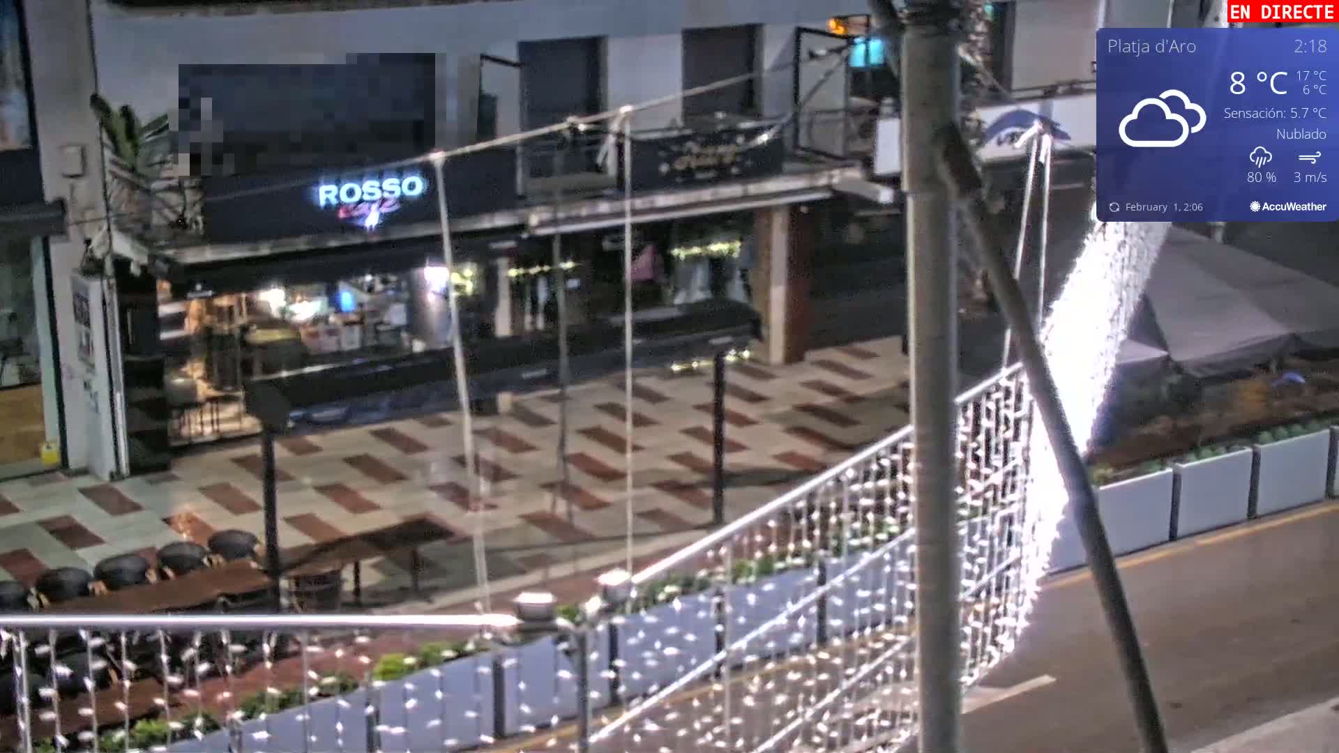 A nighttime, cloudy outdoor scene shows a street with a restaurant, outdoor seating, and string lights along a railing.