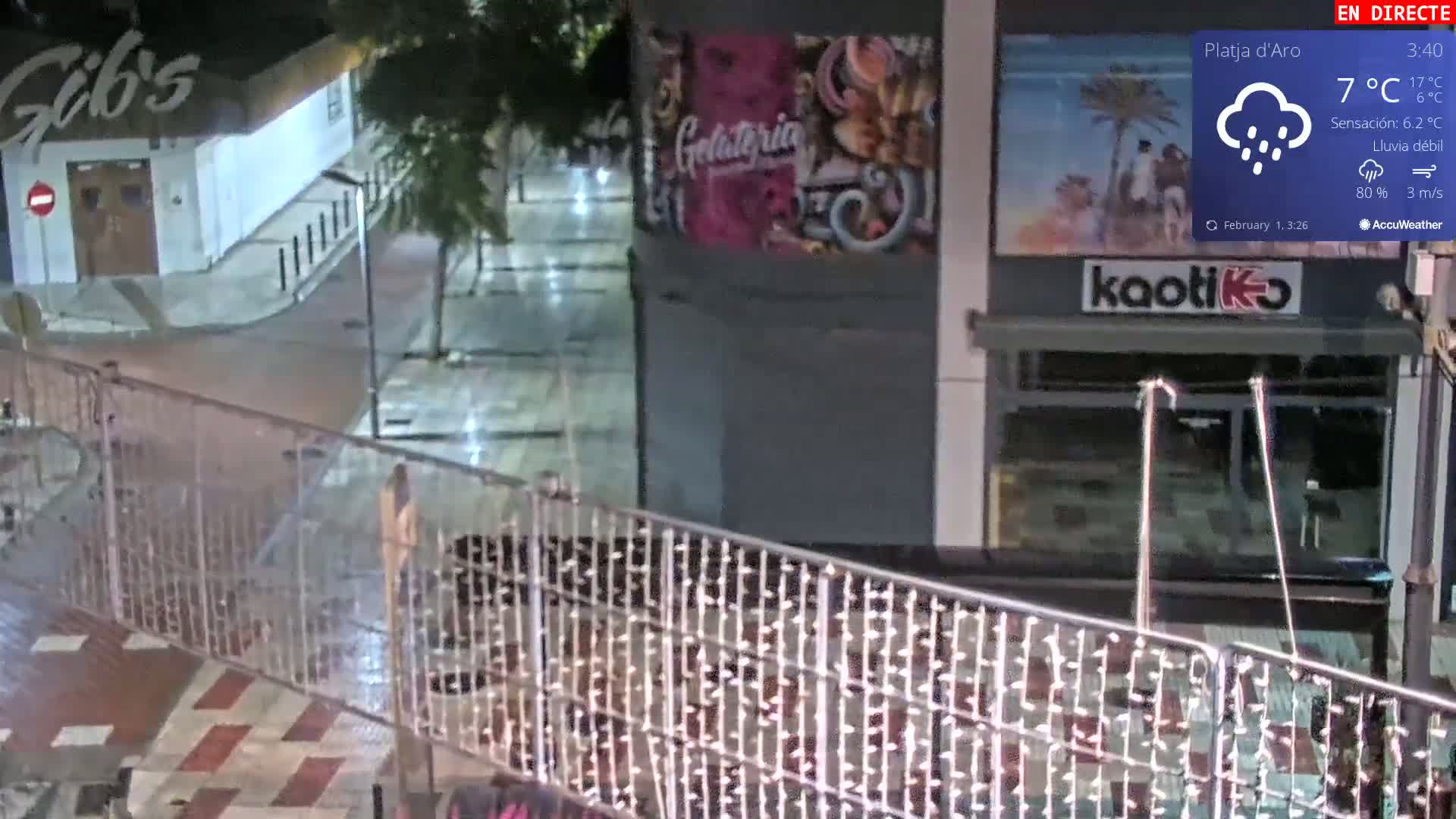 A nighttime street scene shows light rain falling on a wet, paved area with buildings and a metal fence, and a person standing near the fence.
