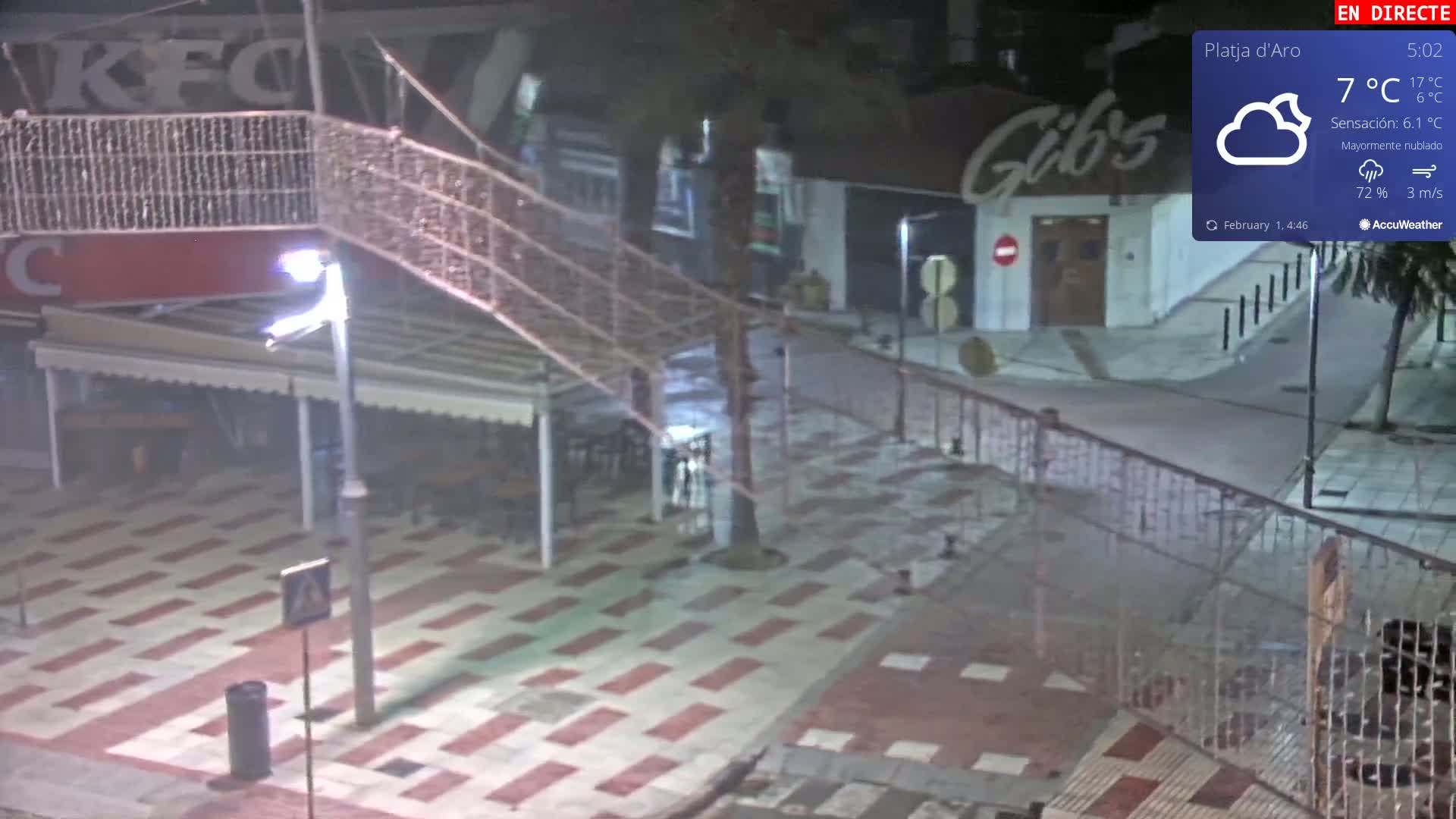 A mostly cloudy night scene shows an empty plaza with a metal walkway, streetlights, and outdoor seating areas under awnings.