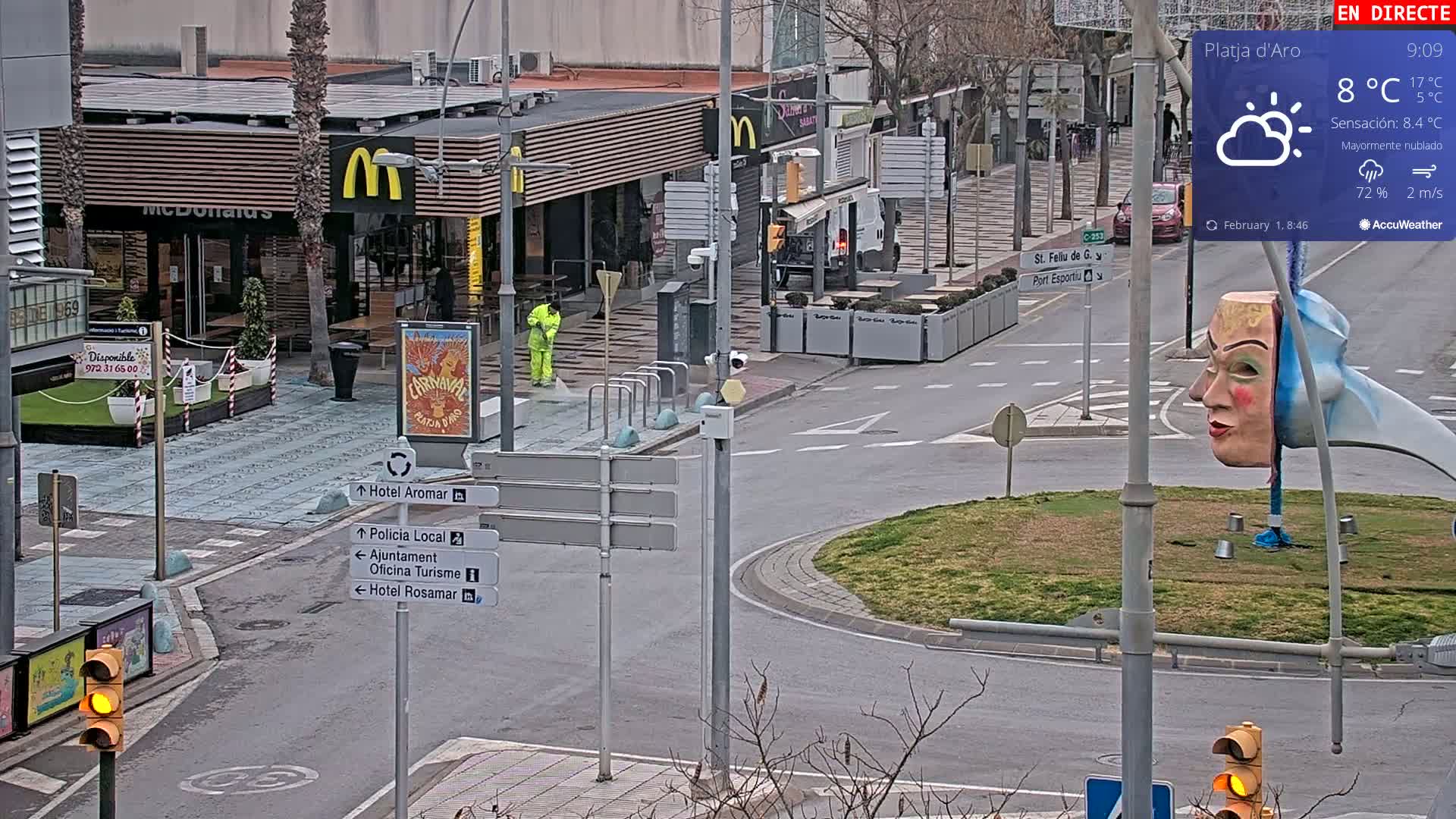 A mostly cloudy day shows a street scene with a McDonald's, street signs, and a person in a yellow jacket, near a roundabout featuring a large carnival mask sculpture.