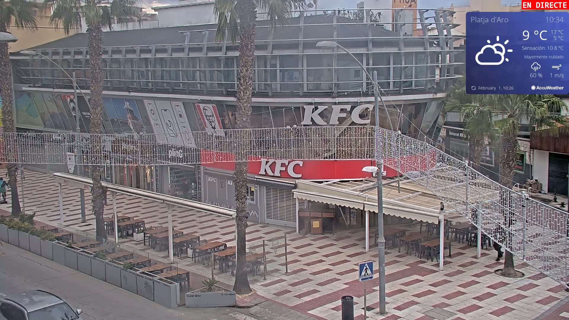 A mostly cloudy day reveals an outdoor KFC restaurant with a patio, surrounded by palm trees and a tiled plaza.