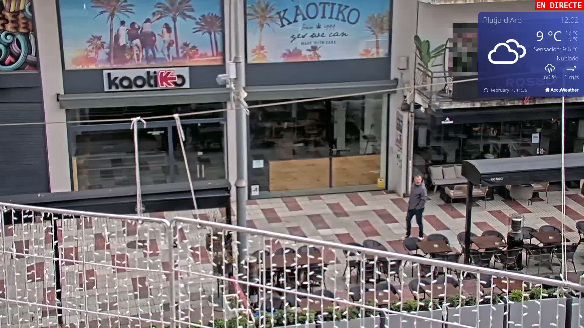 A cloudy, 9°C day shows an outdoor cafe with empty tables and chairs, a man walking by, and a storefront with a beach-themed advertisement.