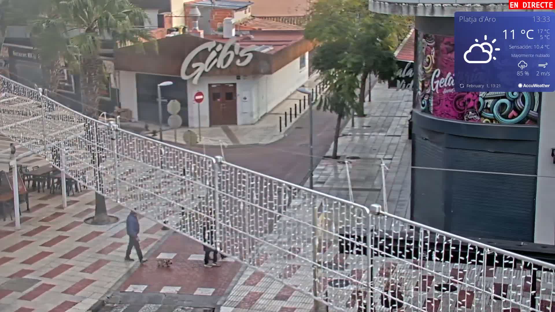 A mostly cloudy outdoor scene shows a man walking a small dog on a paved pedestrian area with string lights overhead, passing in front of a restaurant.