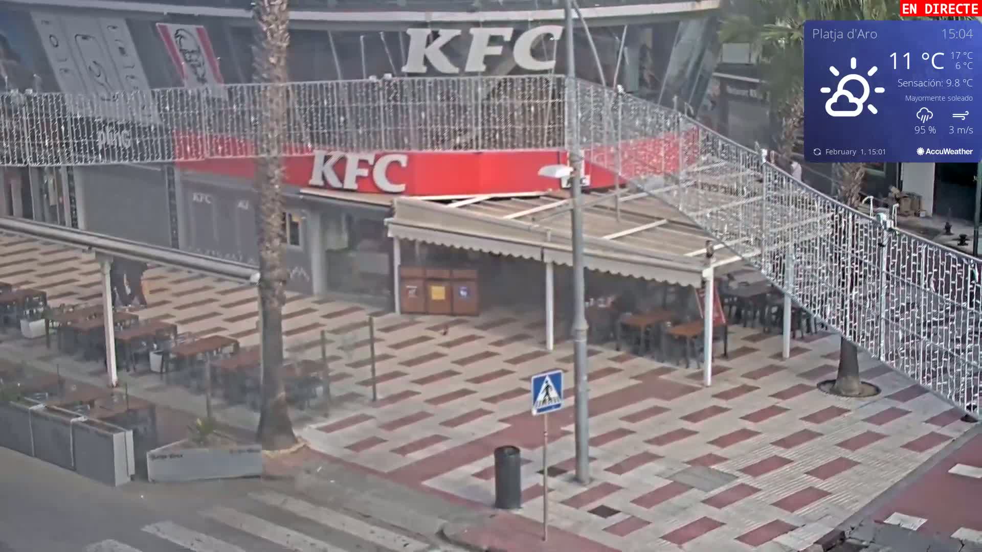 An outdoor plaza with mostly empty tables and chairs in front of a KFC restaurant under partly sunny skies.