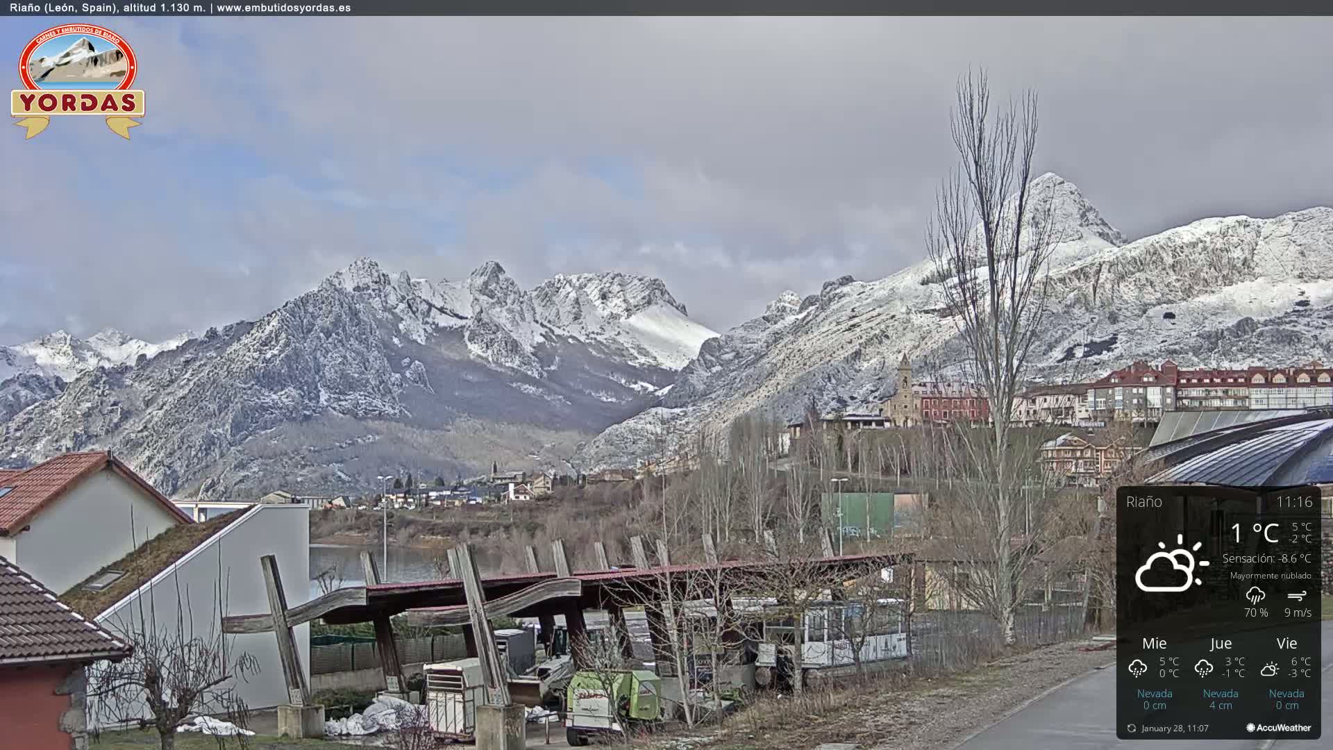 A partly cloudy day reveals a snow-dusted mountain range overlooking a small town and a body of water.