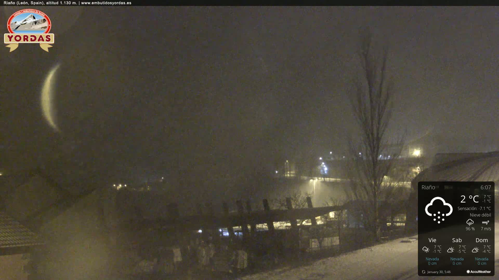 A crescent moon is visible in a nighttime scene of light snowfall over a snow-covered landscape with some buildings and trees.