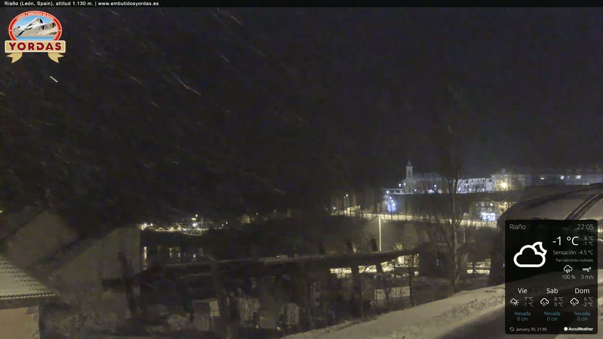 A nighttime snow scene shows a town partially illuminated, with snow falling under a partly cloudy sky.