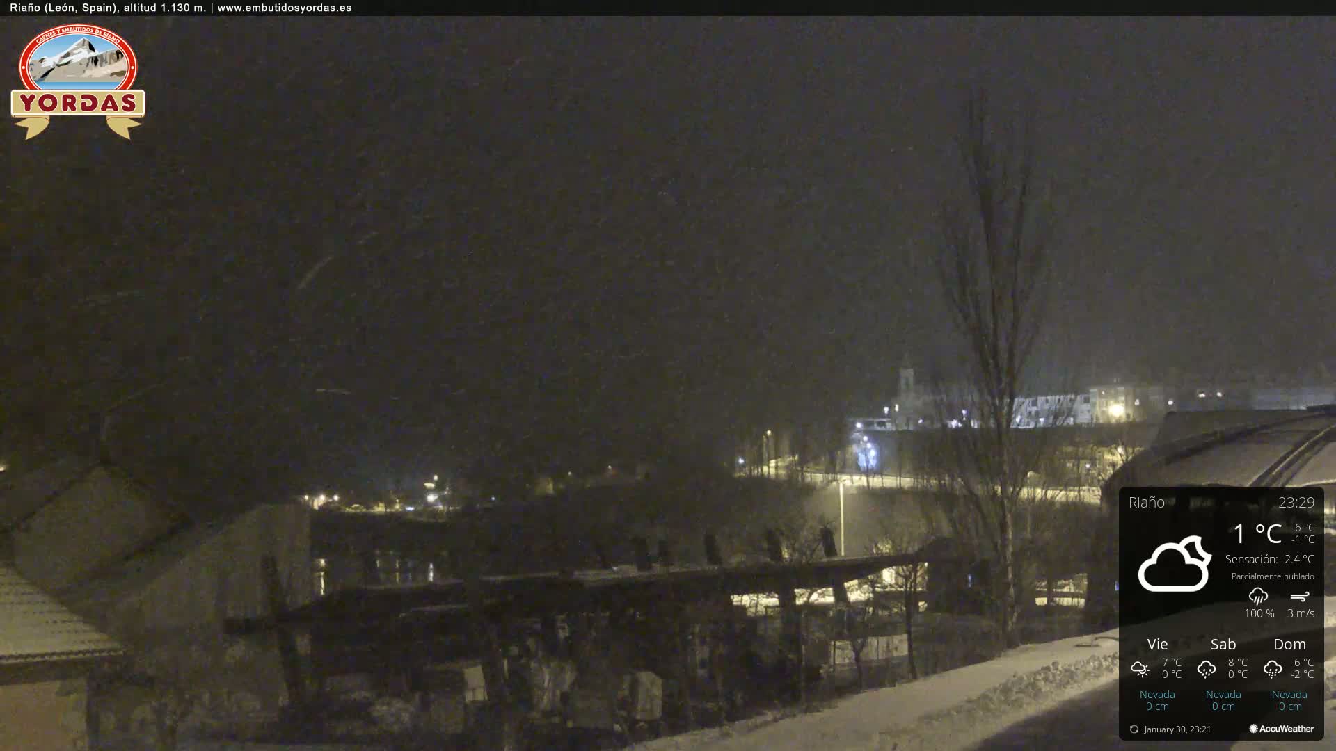 A nighttime scene of a snow-covered town under a partly cloudy sky with light snowfall.