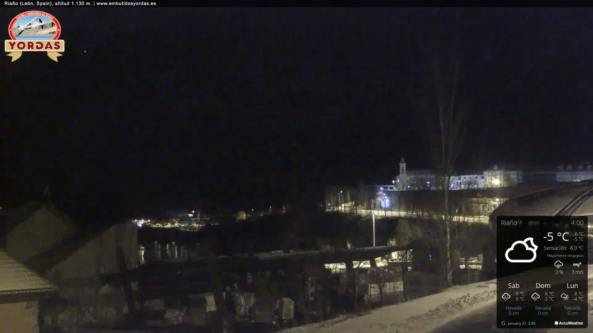 A nighttime view of a snow-covered town under a mostly clear sky, with a few lights visible and a temperature of -5°C.