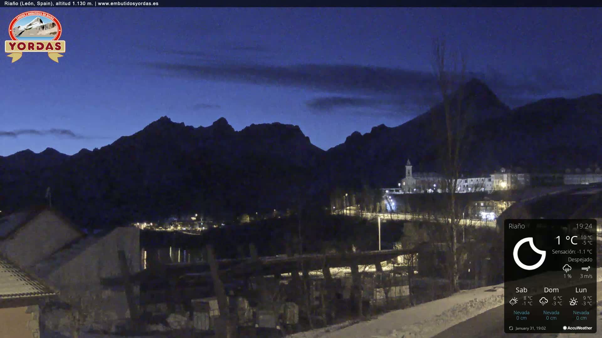 A nighttime view of a snow-dusted town nestled in a valley between dark mountains under a clear, twilight sky.