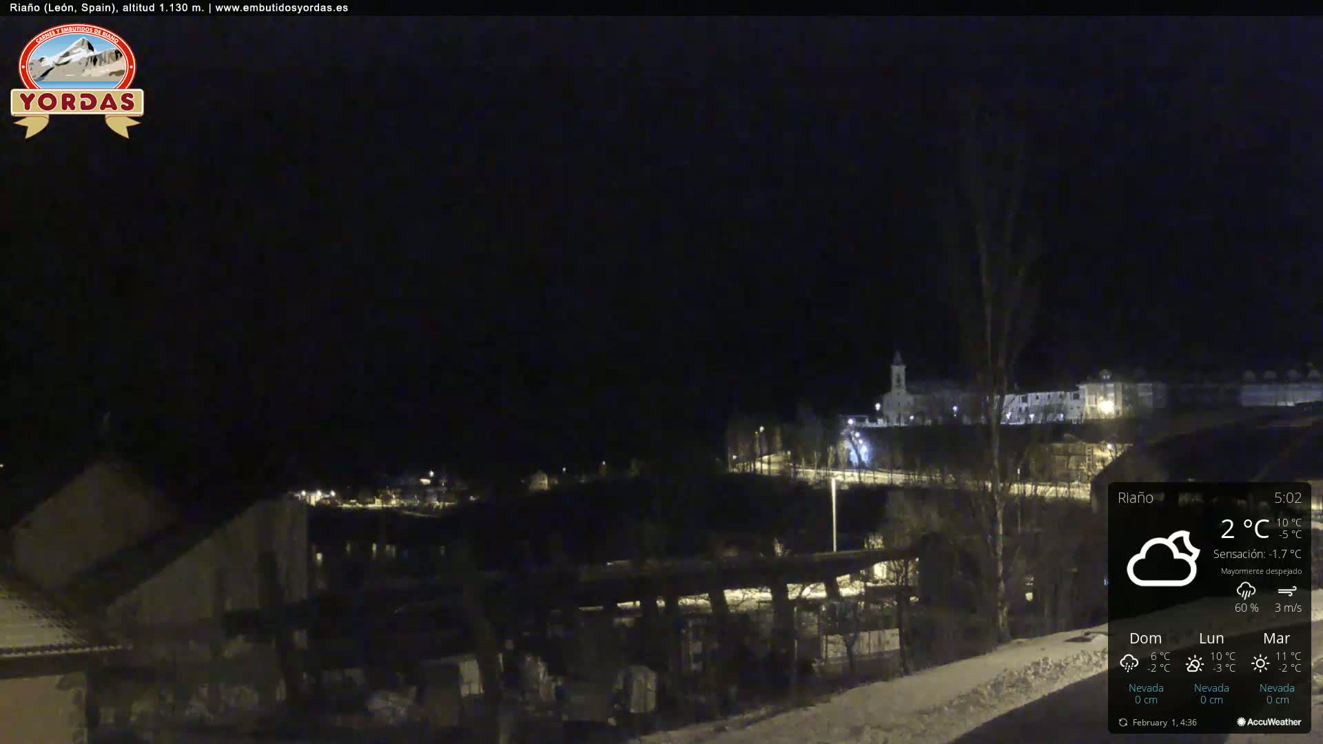 A nighttime view of a snow-covered town, partially illuminated by streetlights, under a mostly clear sky.