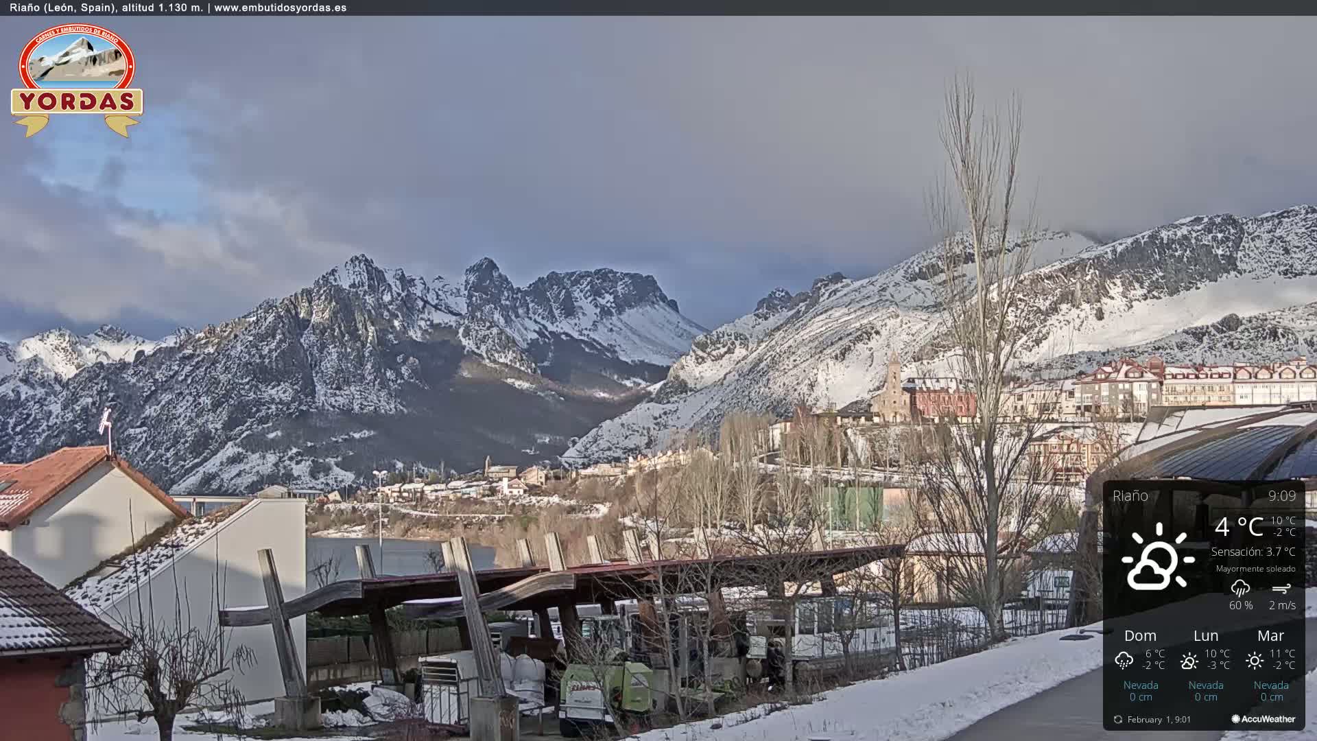 A snow-covered village nestled in a valley between snow-capped mountains under a mostly sunny sky.