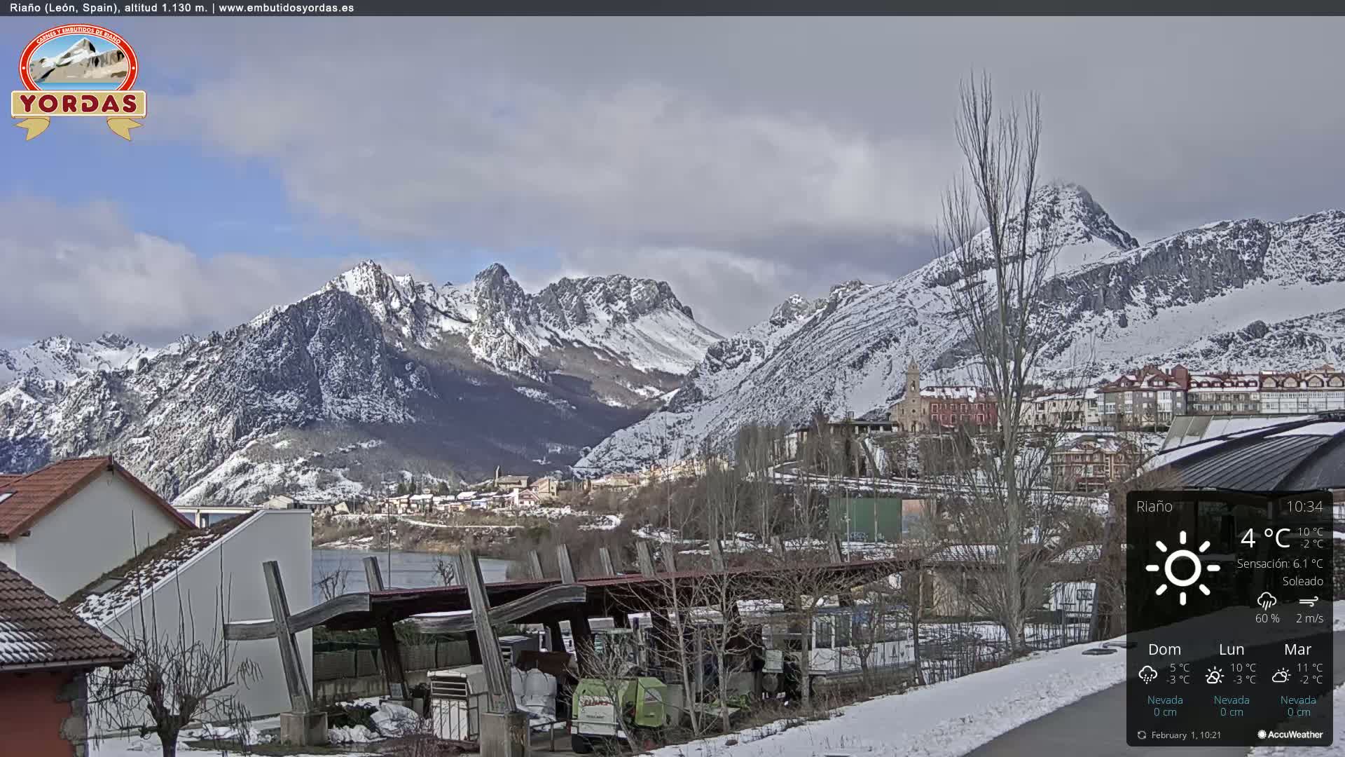 A snow-covered mountain range overlooks a town nestled in a valley, under a partly cloudy sky.