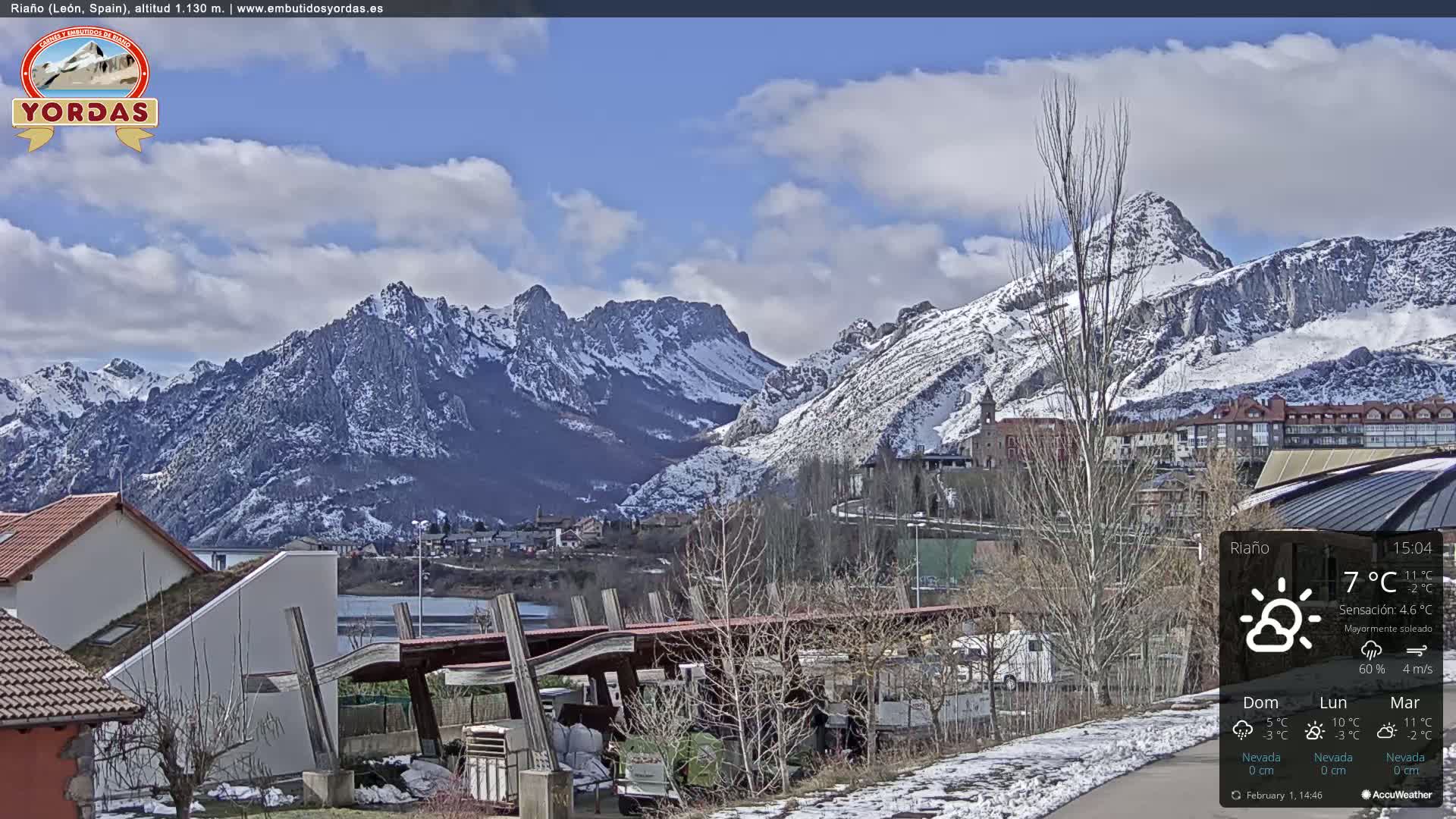 A partly sunny day shows snow-capped mountains in the background overlooking a town with some snow on the ground.