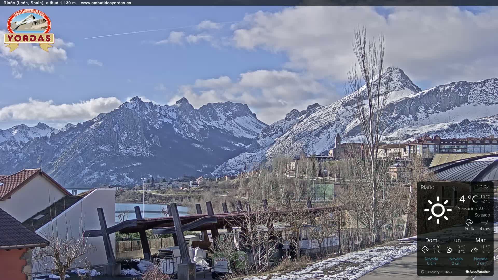 A partly cloudy day reveals snow-capped mountains behind a town nestled beside a lake, with some snow visible on the ground in the foreground.