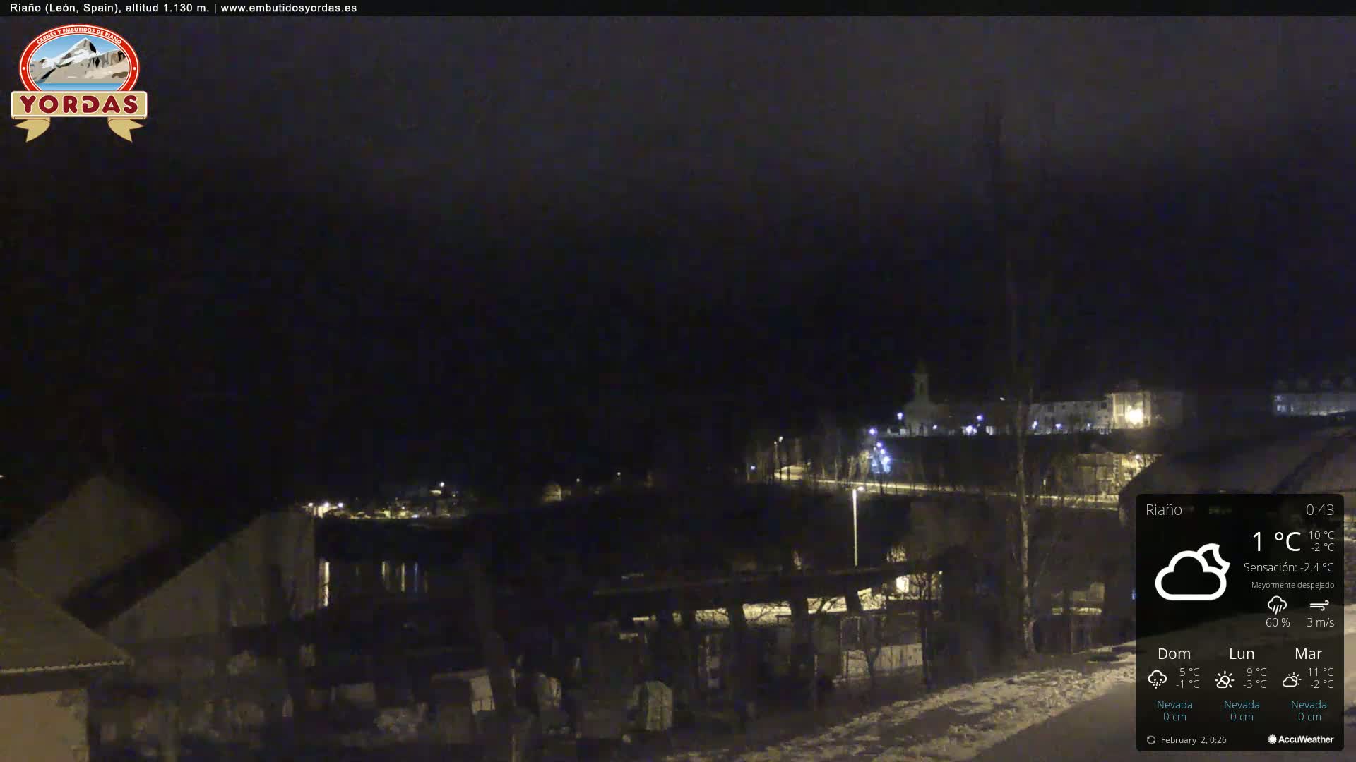 A nighttime view of a small town illuminated by streetlights, with a partly cloudy sky and light snow on the ground.
