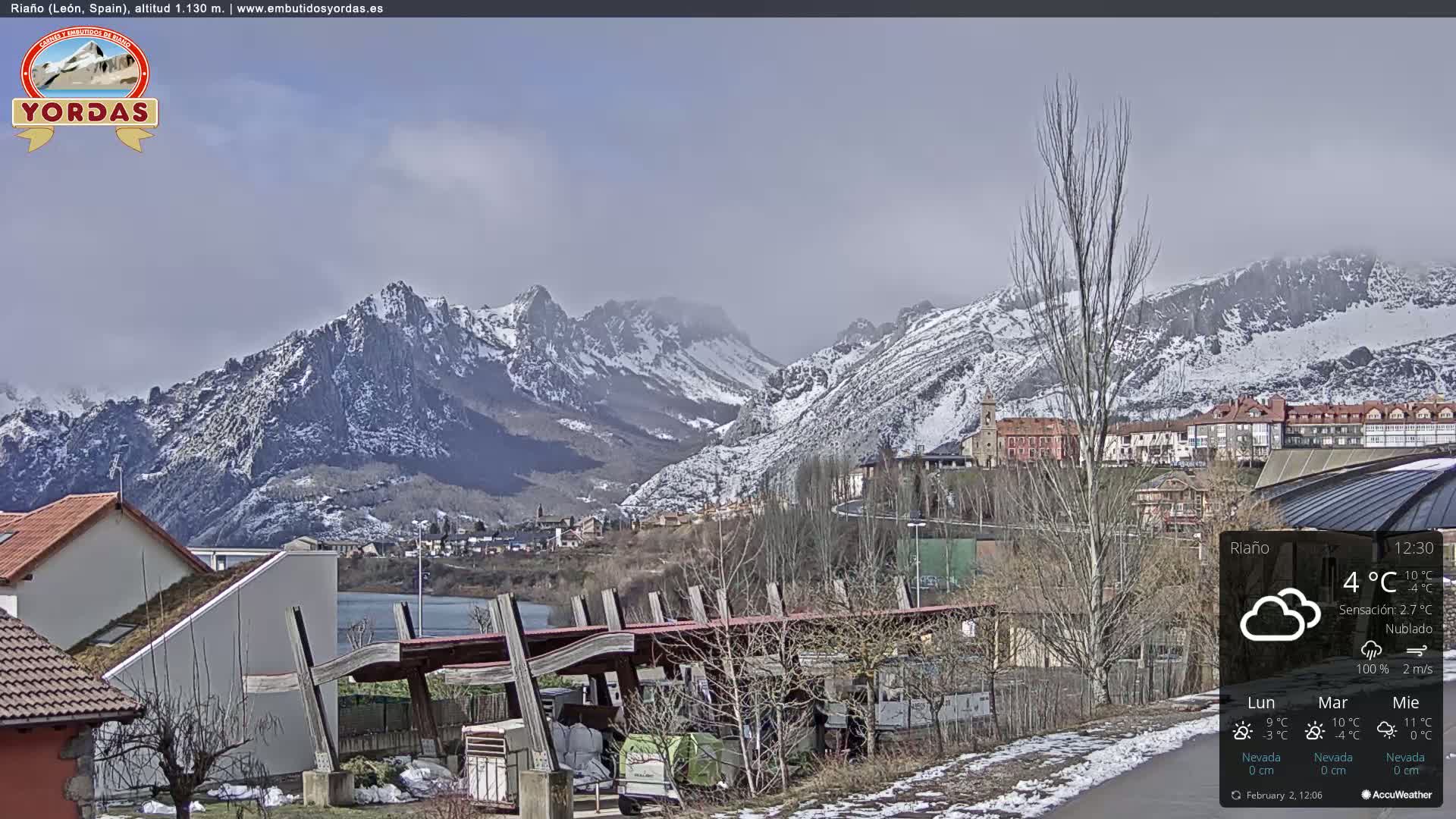 A snow-dusted mountain range overlooks a town nestled beside a lake, with a partly cloudy sky and light snowfall on the ground.