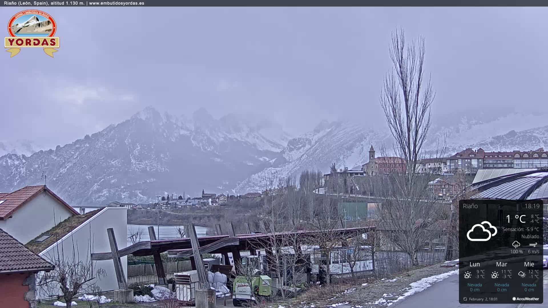 A cloudy, cold day shows snow-dusted mountains in the background, overlooking a town with buildings and a partially visible body of water in the foreground.