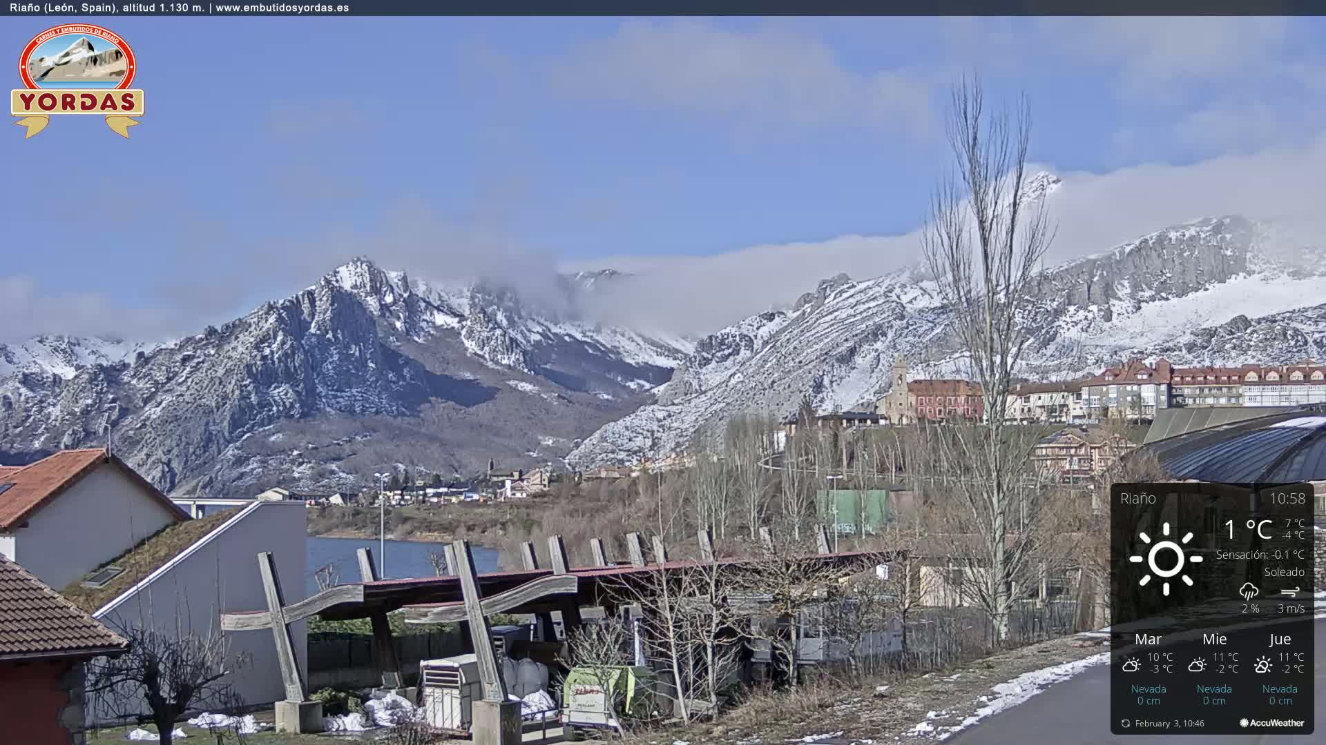 A partly cloudy day reveals snow-capped mountains in the background, overlooking a town and a reservoir with some buildings and structures in the foreground.
