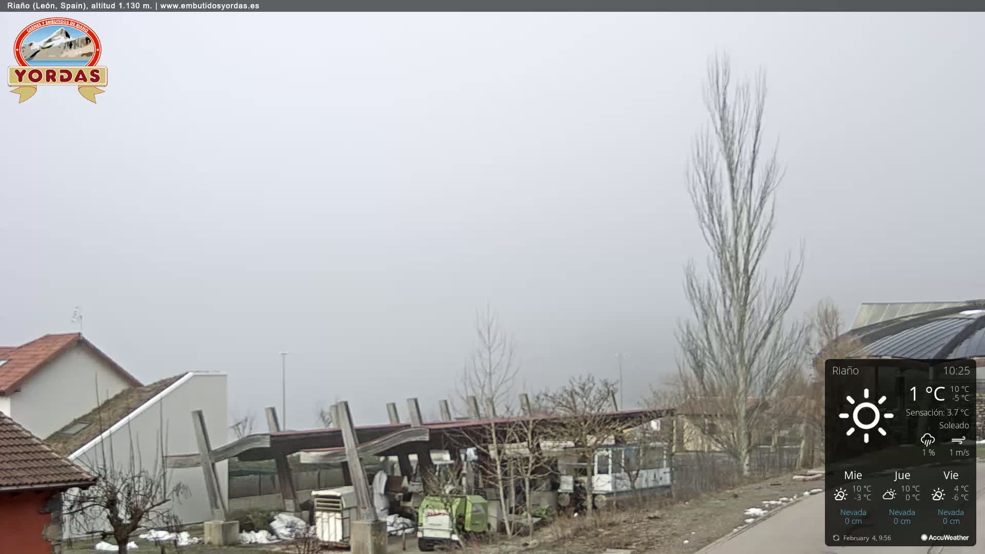 A foggy scene shows several buildings, a few bare trees, and some machinery in a yard.