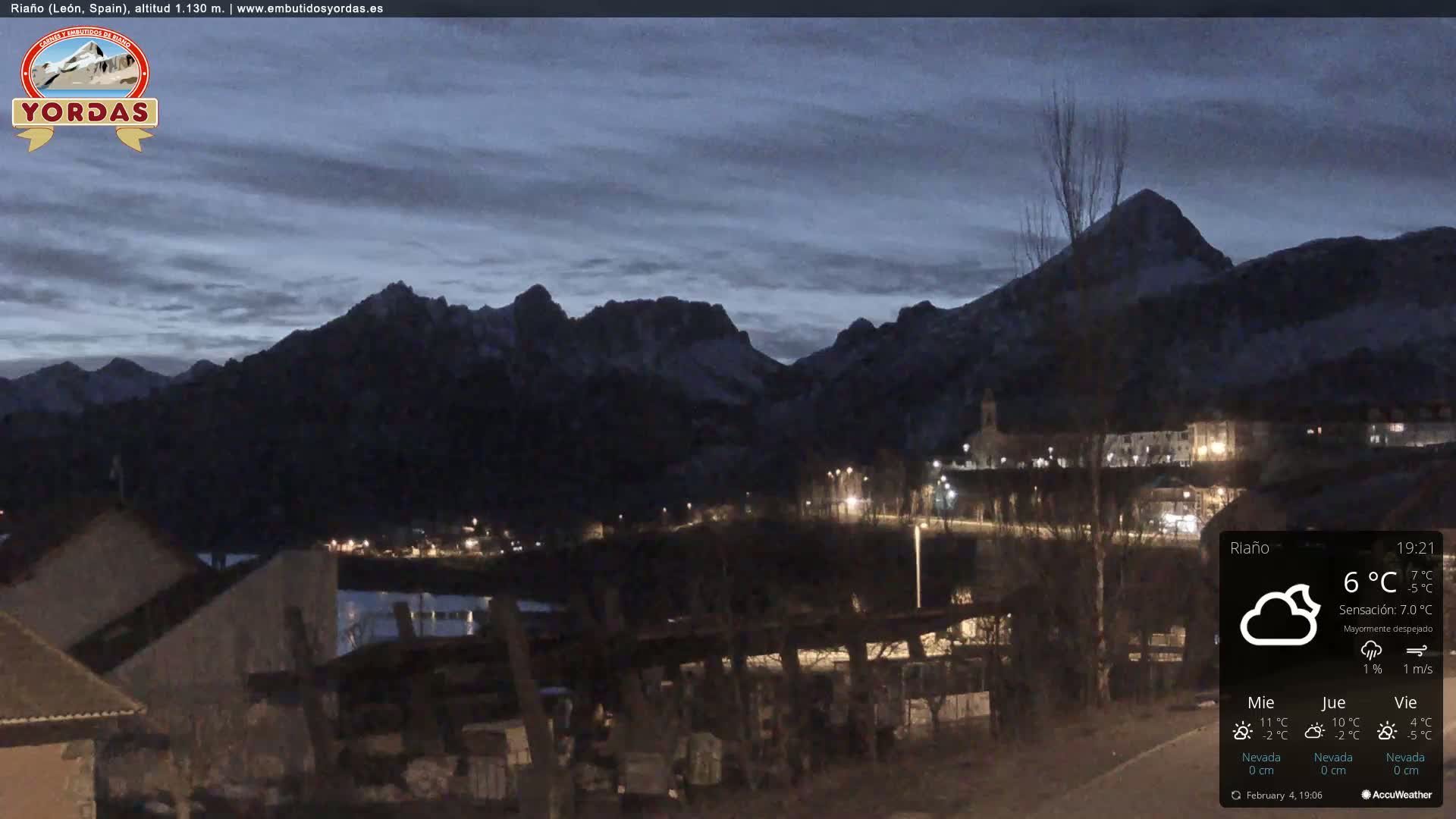 A nighttime view of a village nestled at the base of snow-capped mountains under a mostly clear sky.