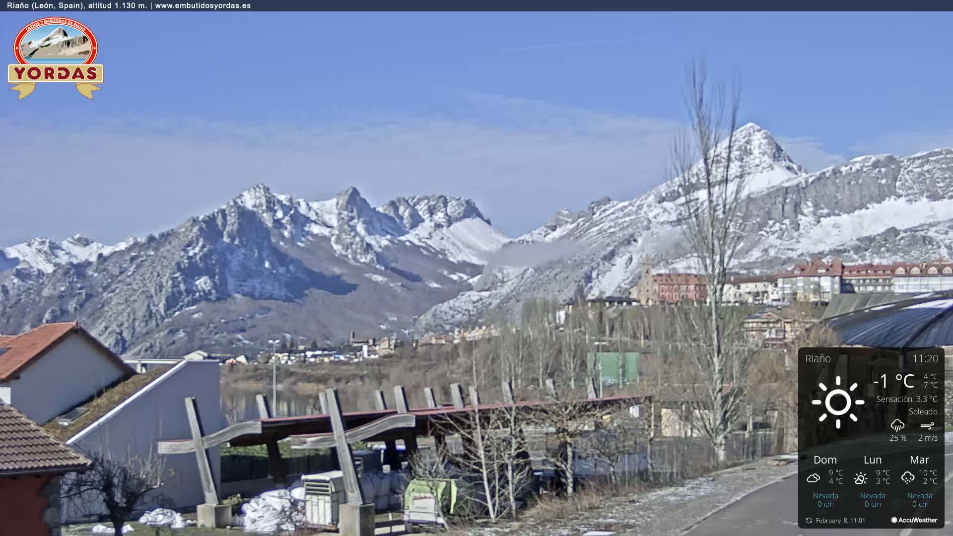 A sunny, cold day shows a snow-capped mountain range behind a town with buildings and a covered structure in the foreground.