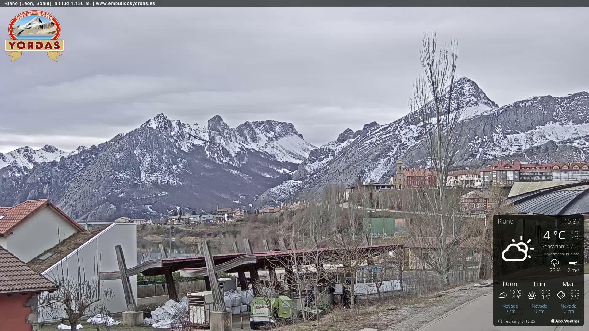 A partly cloudy day reveals snow-dusted mountains behind a town and buildings, with some structures under a covered shelter.
