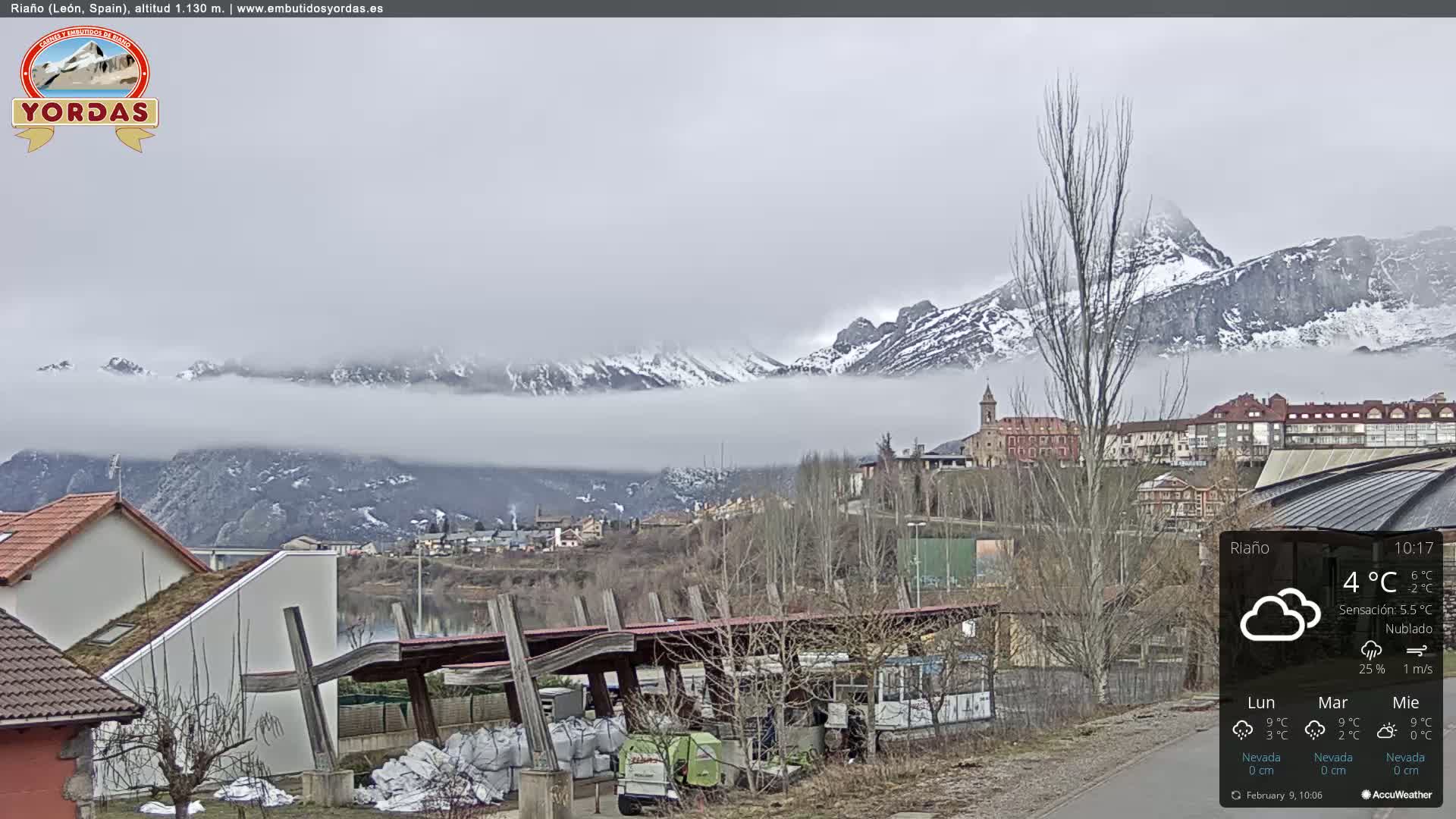 A town nestled in a valley is shrouded in fog, with snow-capped mountains visible in the distance under an overcast sky.
