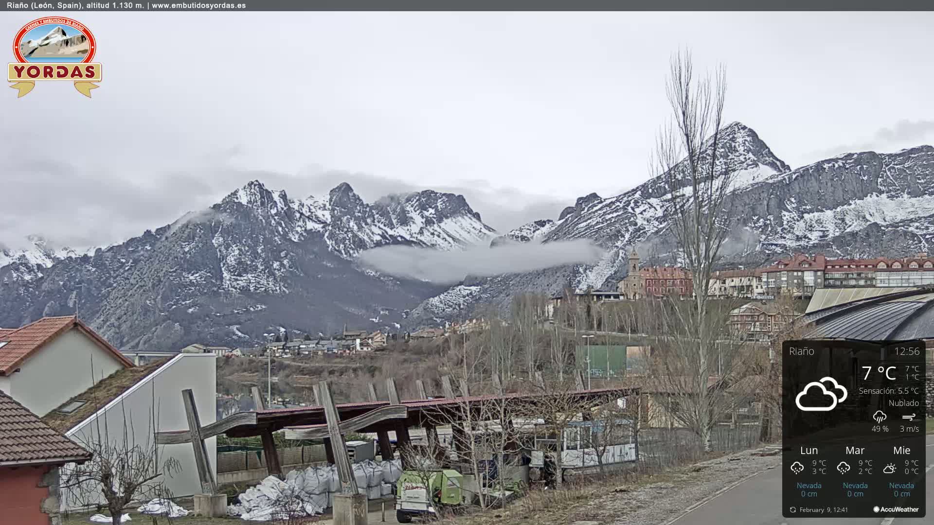 A town nestled in a valley between snow-capped mountains, partially obscured by clouds, under a cloudy sky.