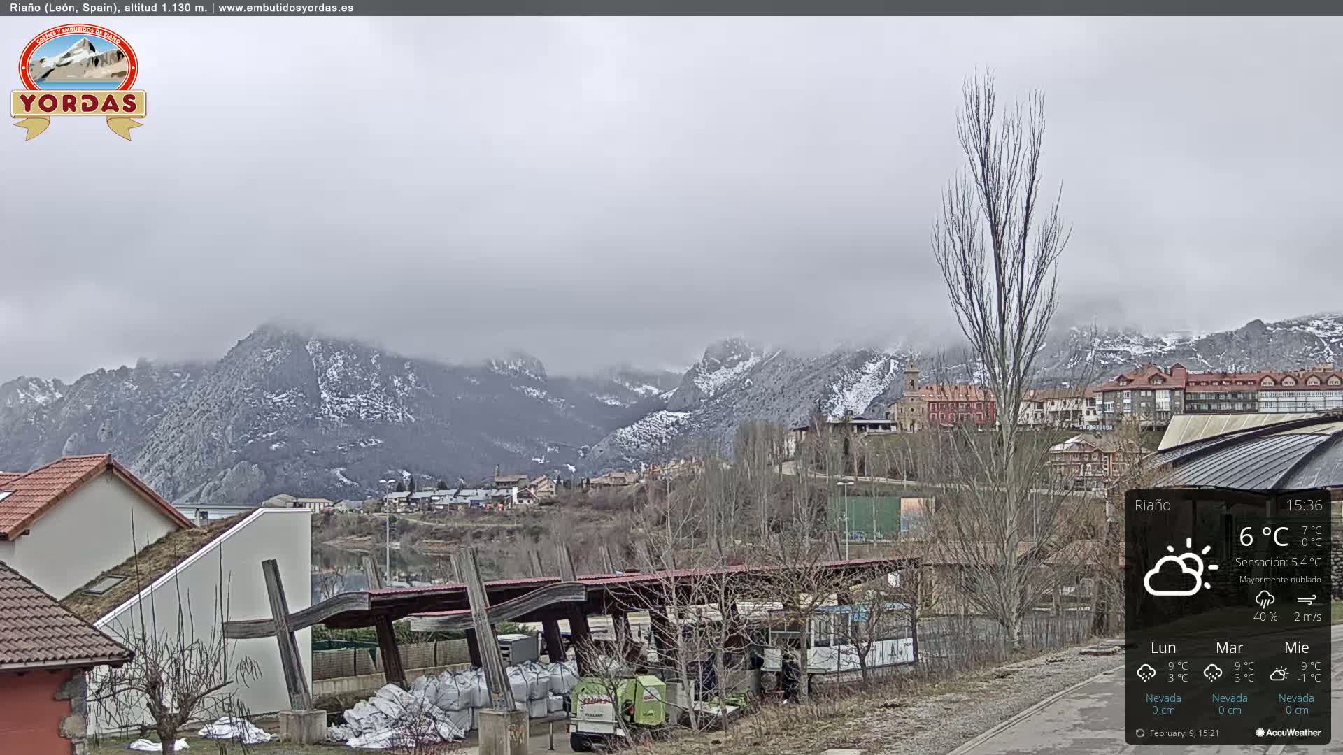 A mostly cloudy day reveals a snow-dusted mountain range overlooking a small town with buildings and a parking area, some of which are partially obscured by fog.