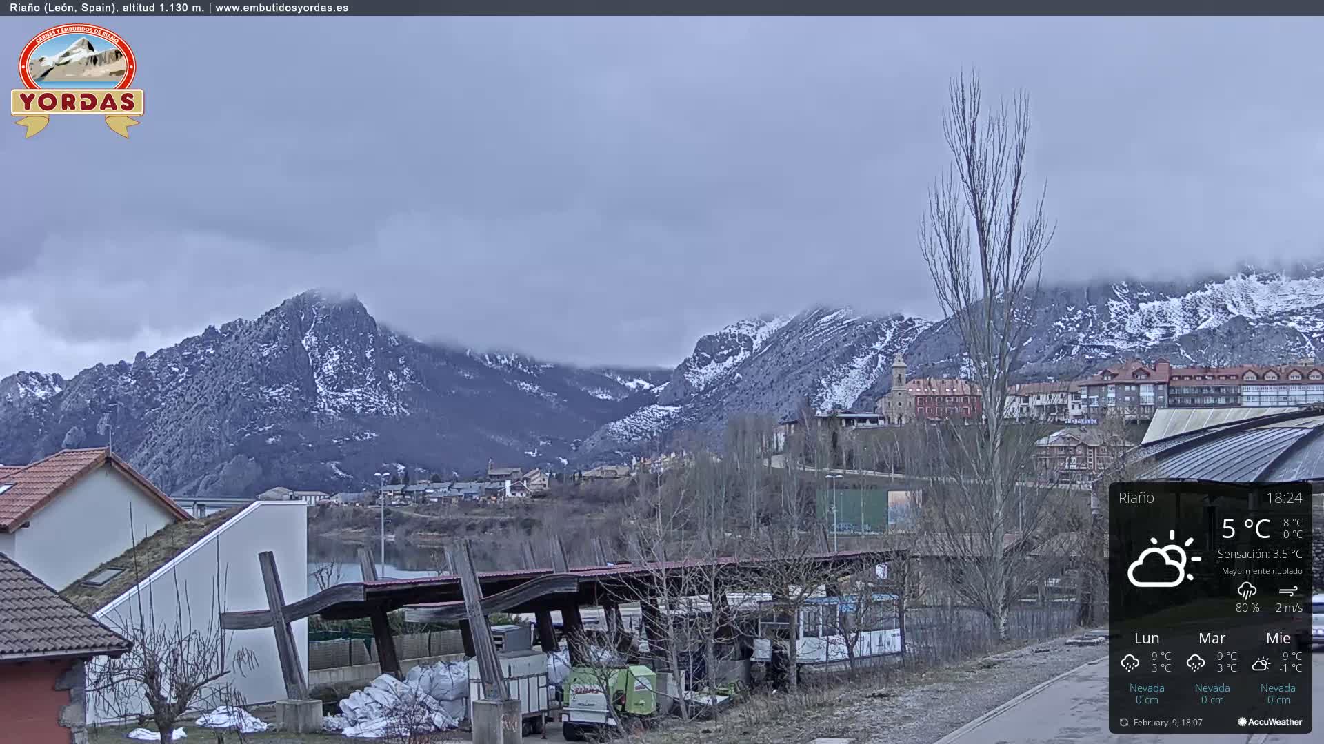 A mostly cloudy view of a snow-dusted mountain range overlooking a small town nestled beside a body of water.