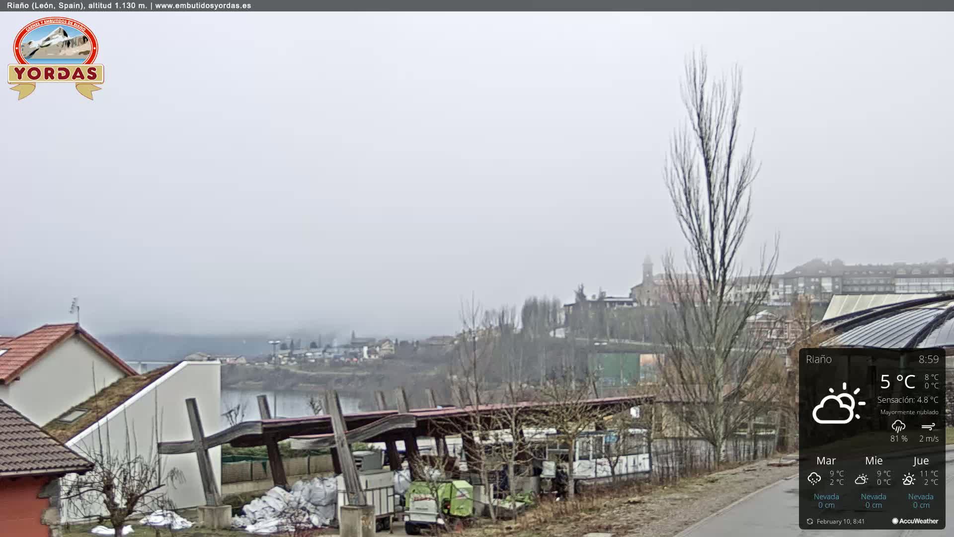 A foggy view of a town by a river, with leafless trees and buildings, including a partially covered structure, in the foreground.