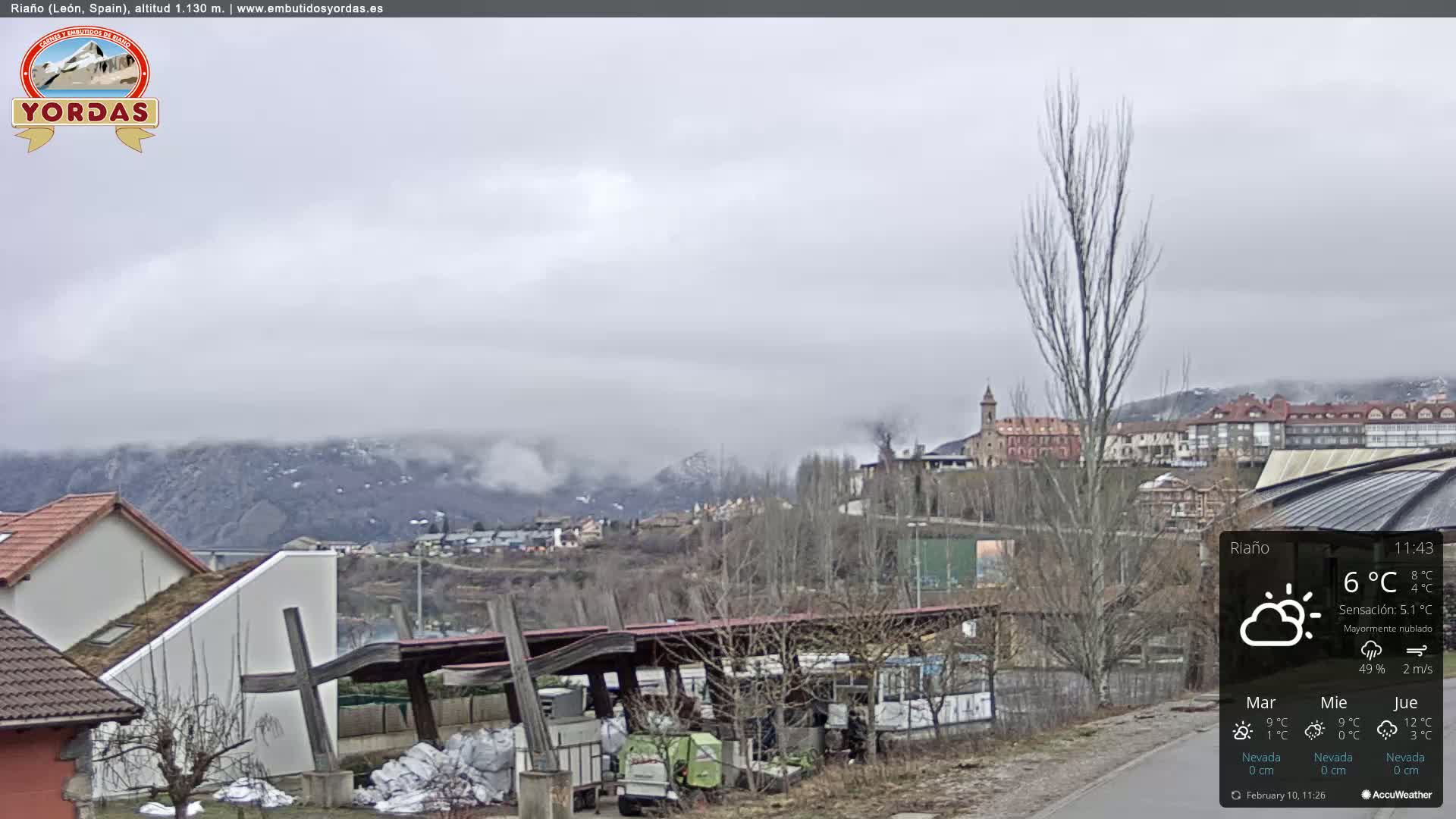 A mostly cloudy scene overlooks a town nestled in a valley, with snow-dusted mountains in the background.
