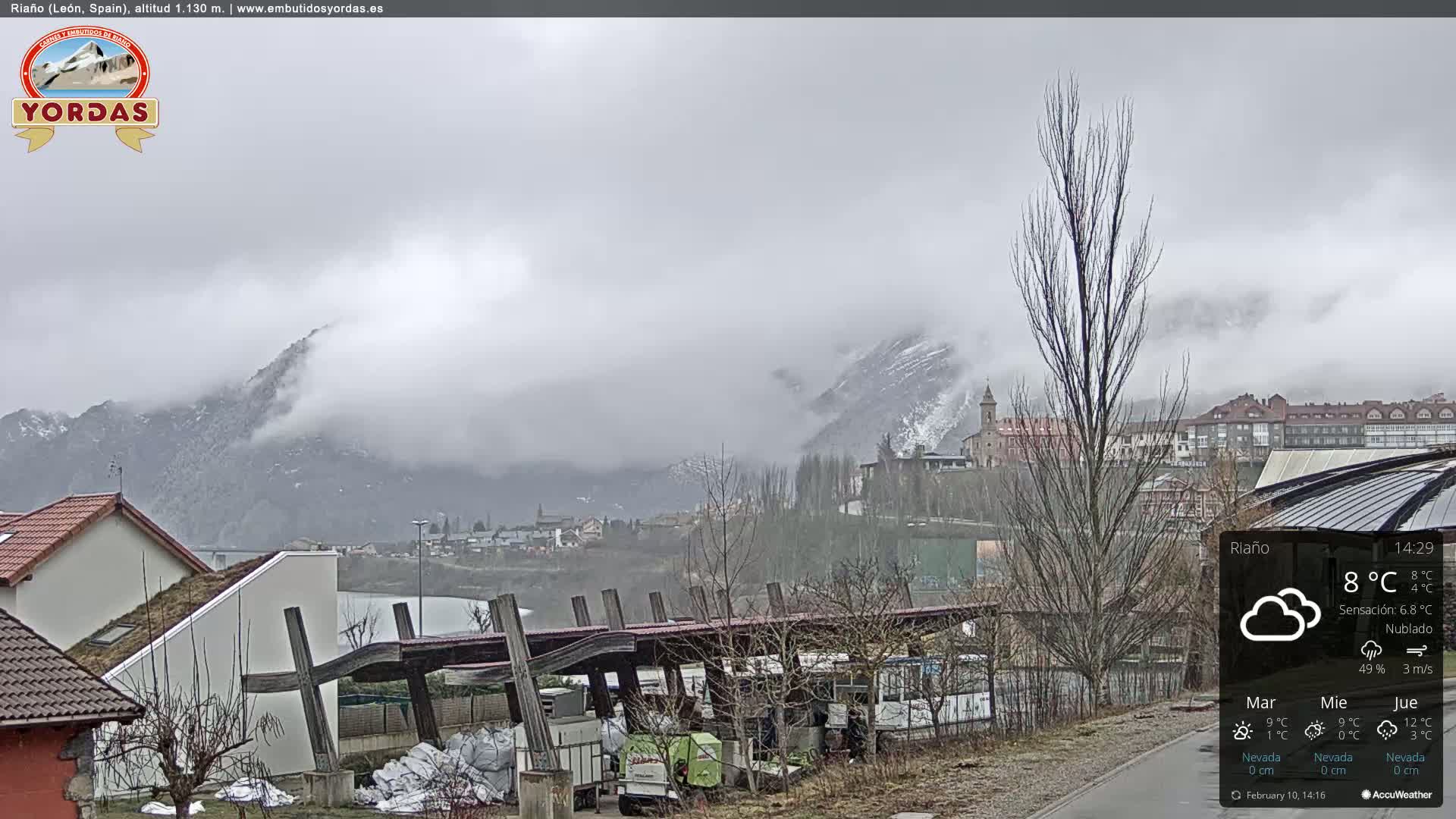 A cloudy, overcast day reveals a town nestled in a valley surrounded by snow-capped mountains partially obscured by fog.