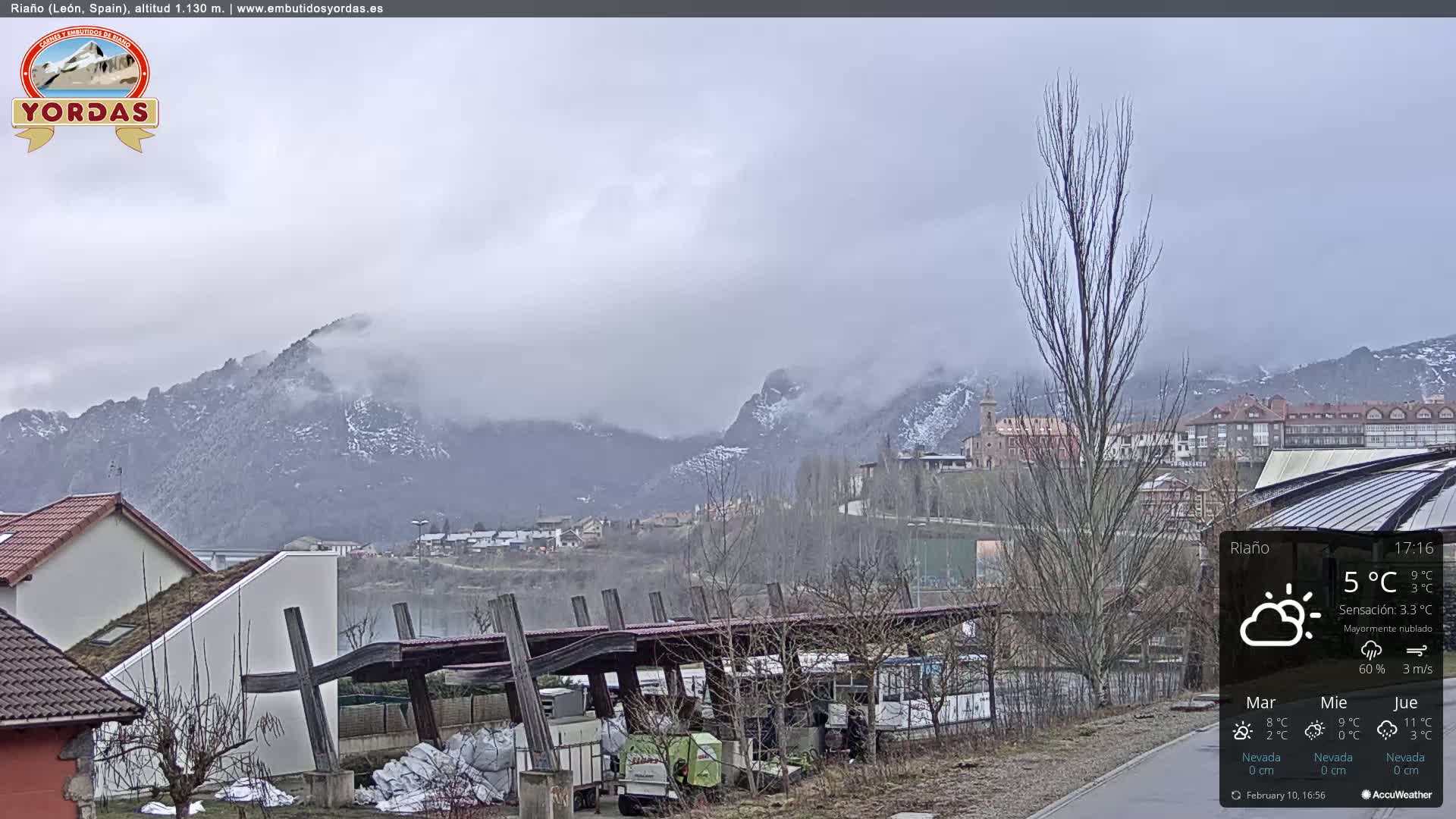 A mostly cloudy day reveals snow-capped mountains in the distance overlooking a town with buildings and a covered structure, all situated near a body of water.