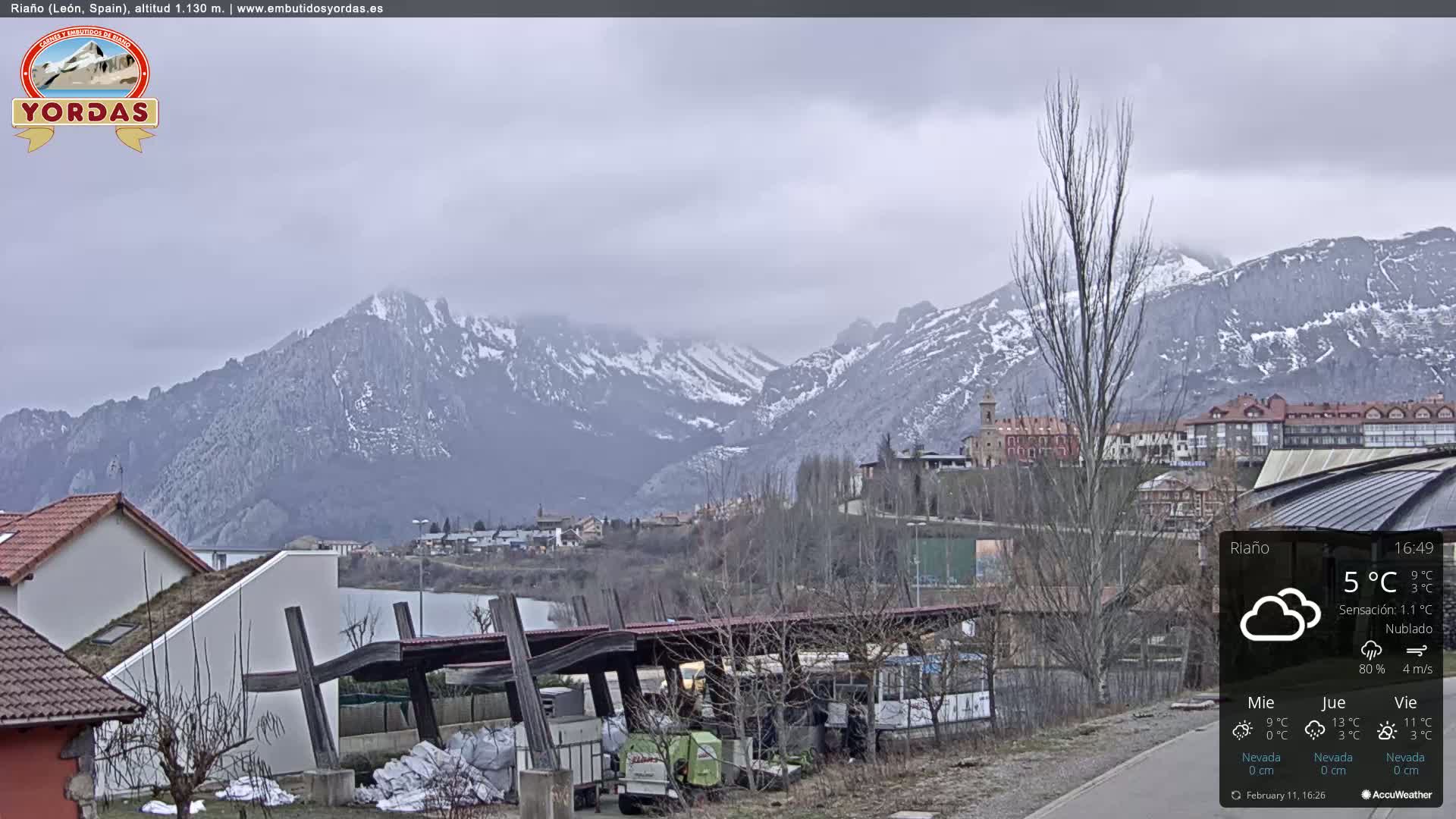 A partly cloudy day reveals snow-capped mountains in the distance, overlooking a town with buildings and a covered structure near a body of water.