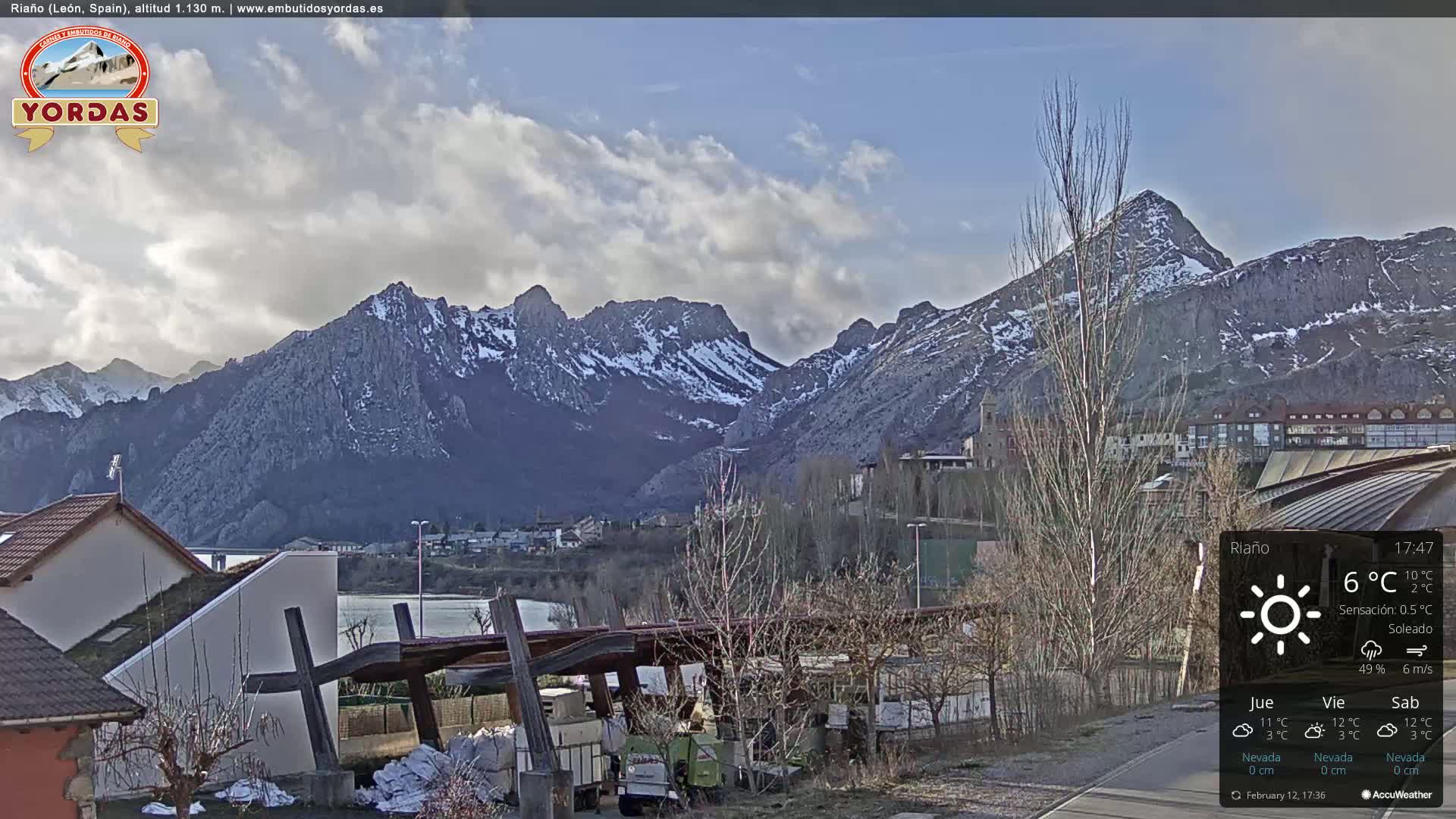 A partly cloudy day reveals snow-capped mountains behind a town nestled in a valley, with a few buildings and some trees in the foreground.