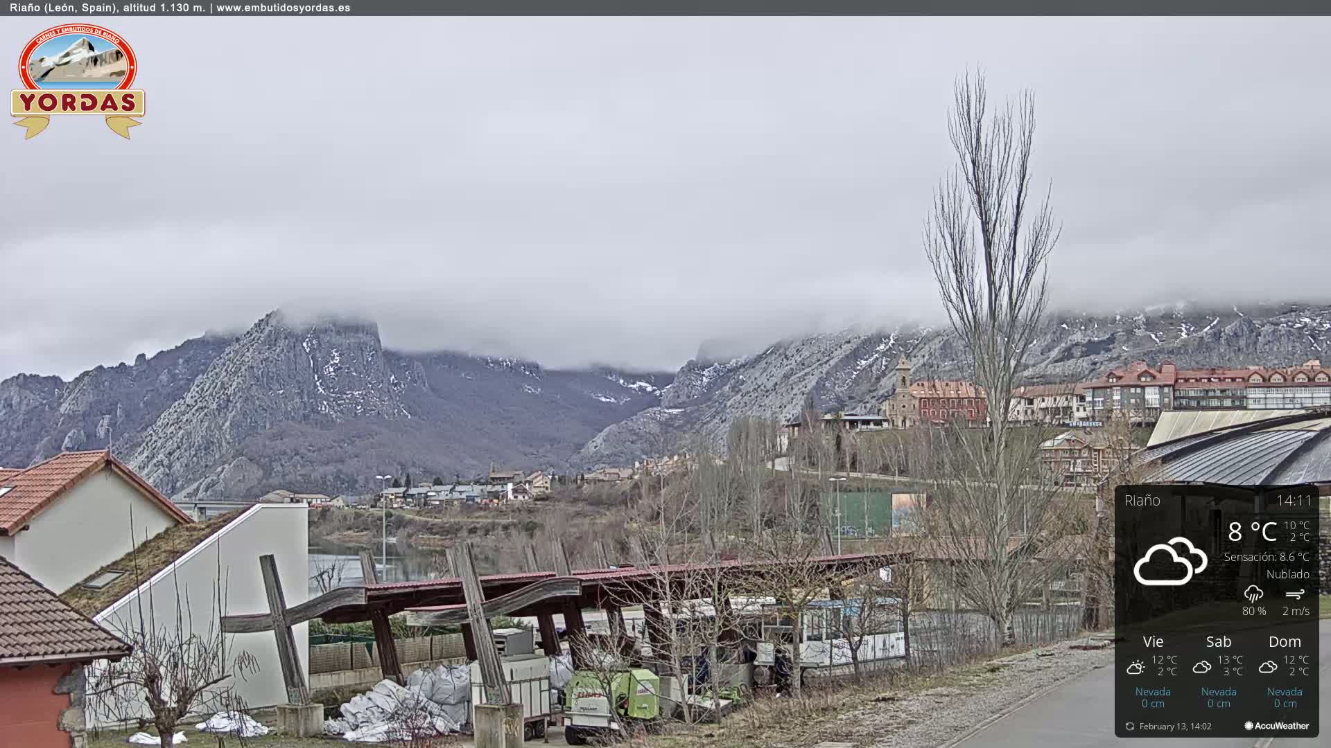 A cloudy day in a mountainous town, with a partially visible river and buildings nestled at the base of snow-dusted peaks.