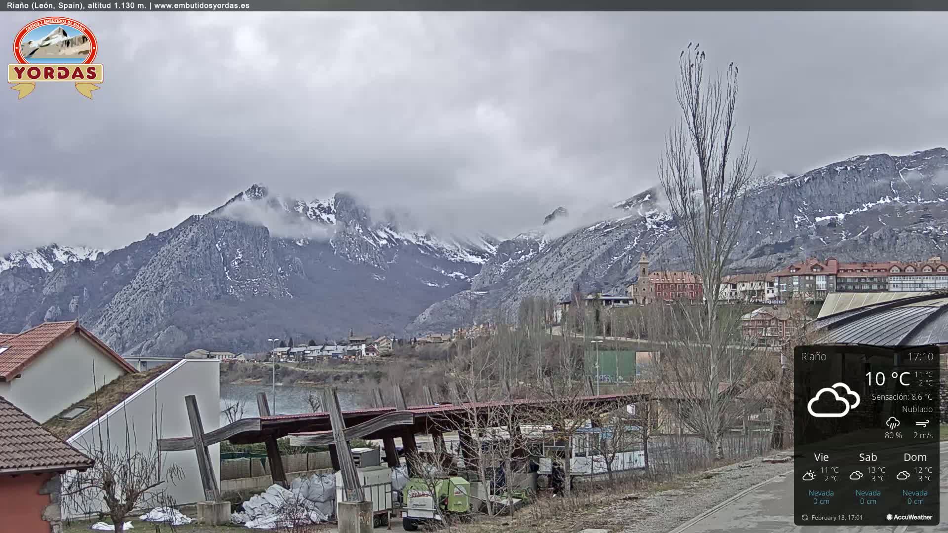 A village nestled in a valley, overlooked by snow-capped mountains under a cloudy sky.