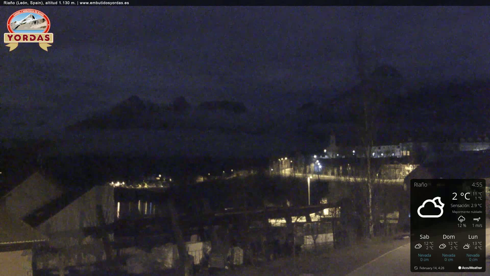 A nighttime view of a town nestled in a valley, partially obscured by clouds and mountains under a mostly cloudy sky.
