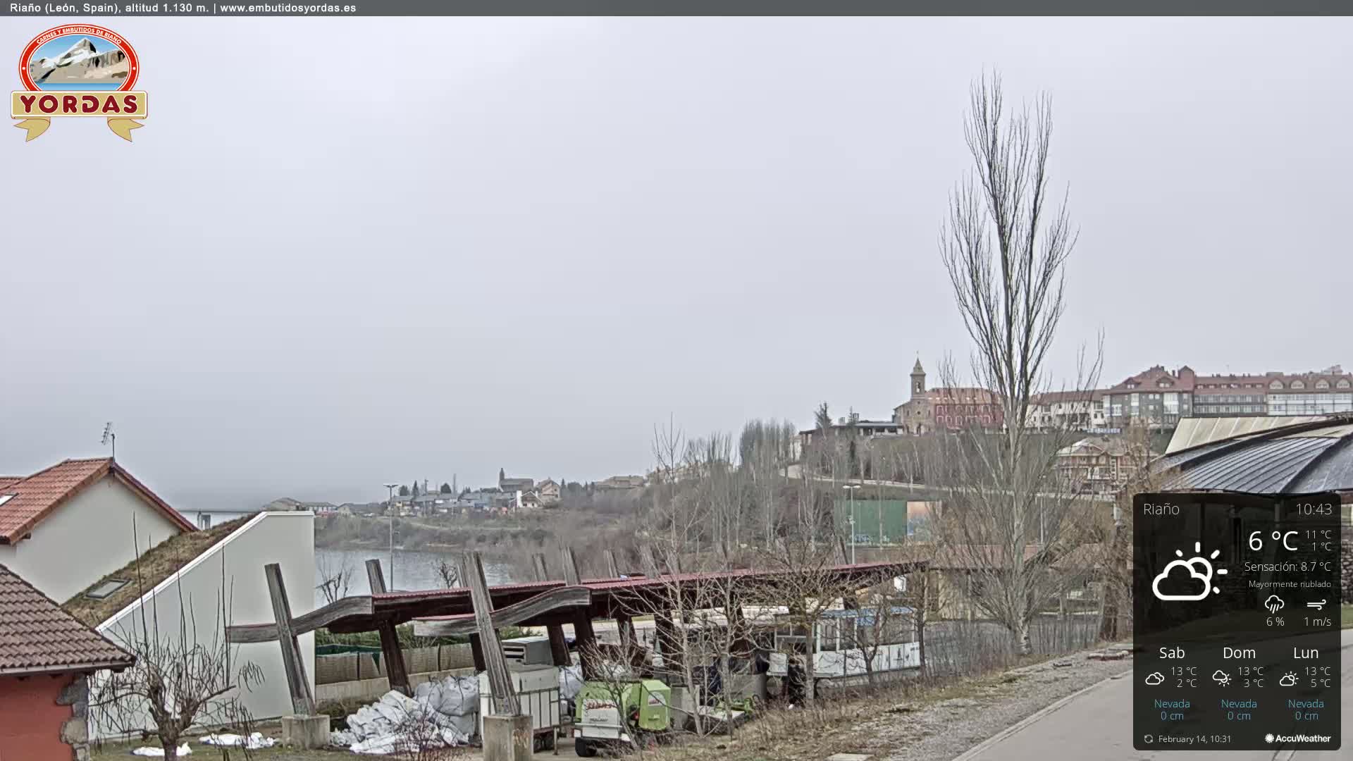 A mostly cloudy day reveals a town nestled beside a body of water, with bare trees and buildings visible, and a covered structure in the foreground.