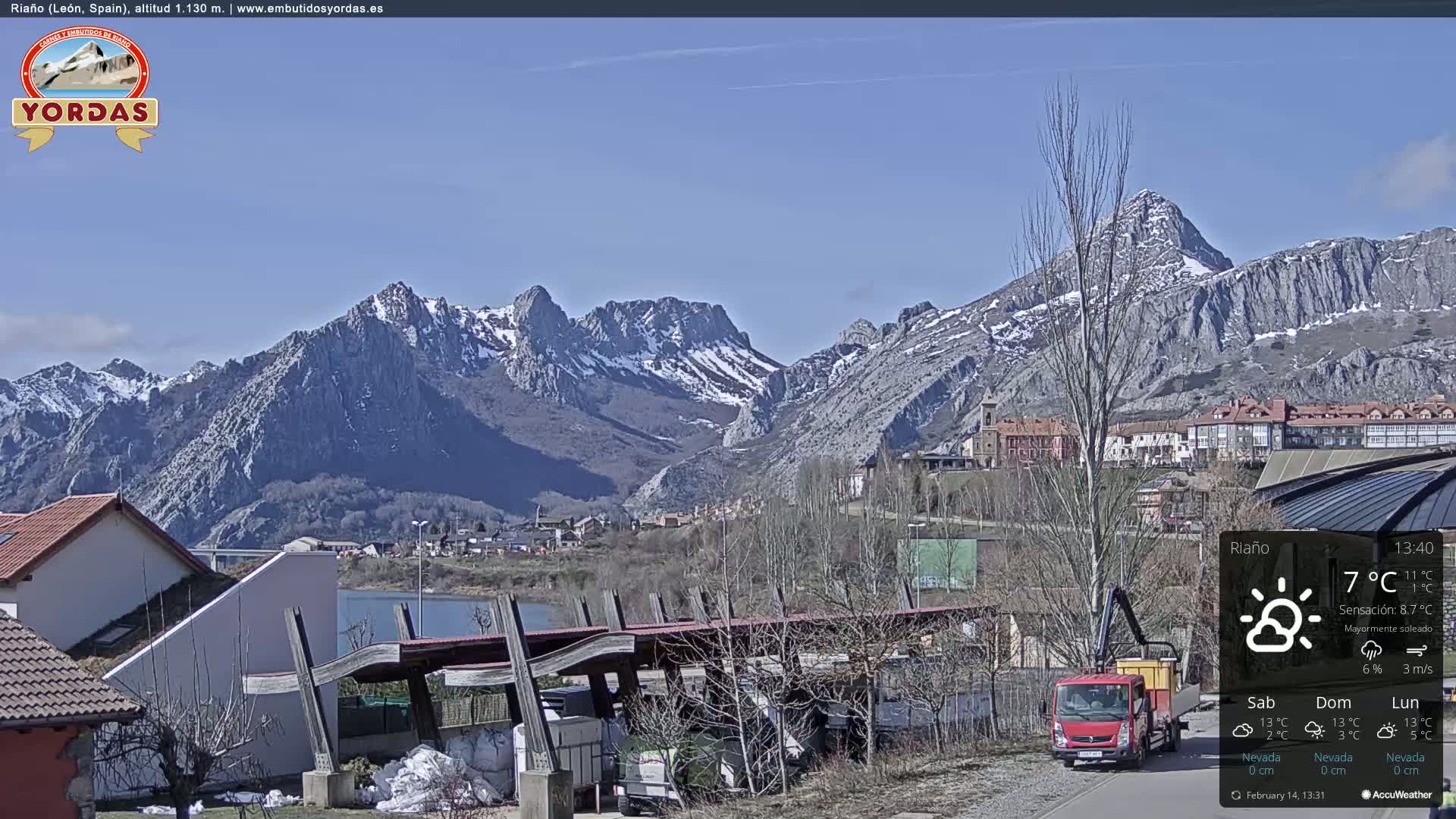 A town nestled beside a lake, with snow-capped mountains in the background under a mostly sunny sky.