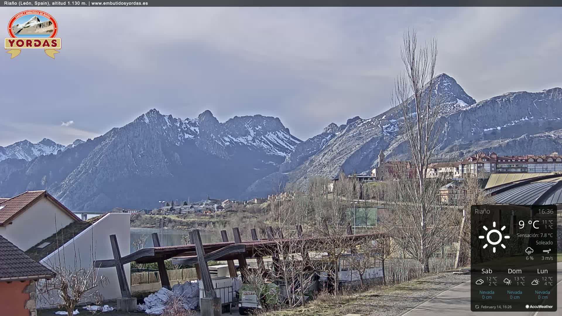 A partly sunny view of a town nestled in a valley, with snow-capped mountains in the background.