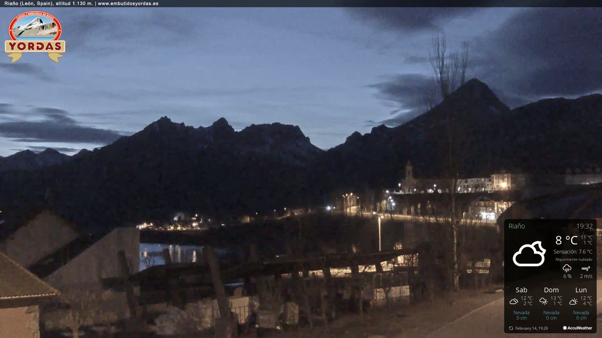 A small town nestled at the base of dark, snow-capped mountains under a mostly cloudy twilight sky.