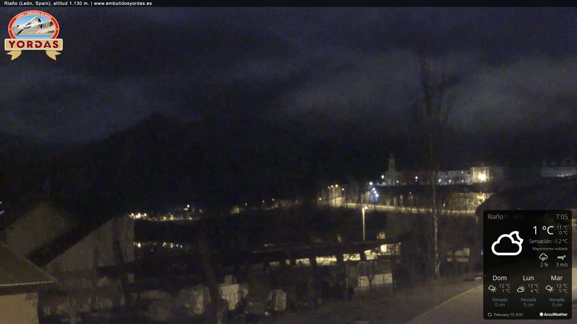A nighttime view of a town nestled beneath a mostly cloudy sky, with faint lights illuminating the buildings and a mountain in the distance.