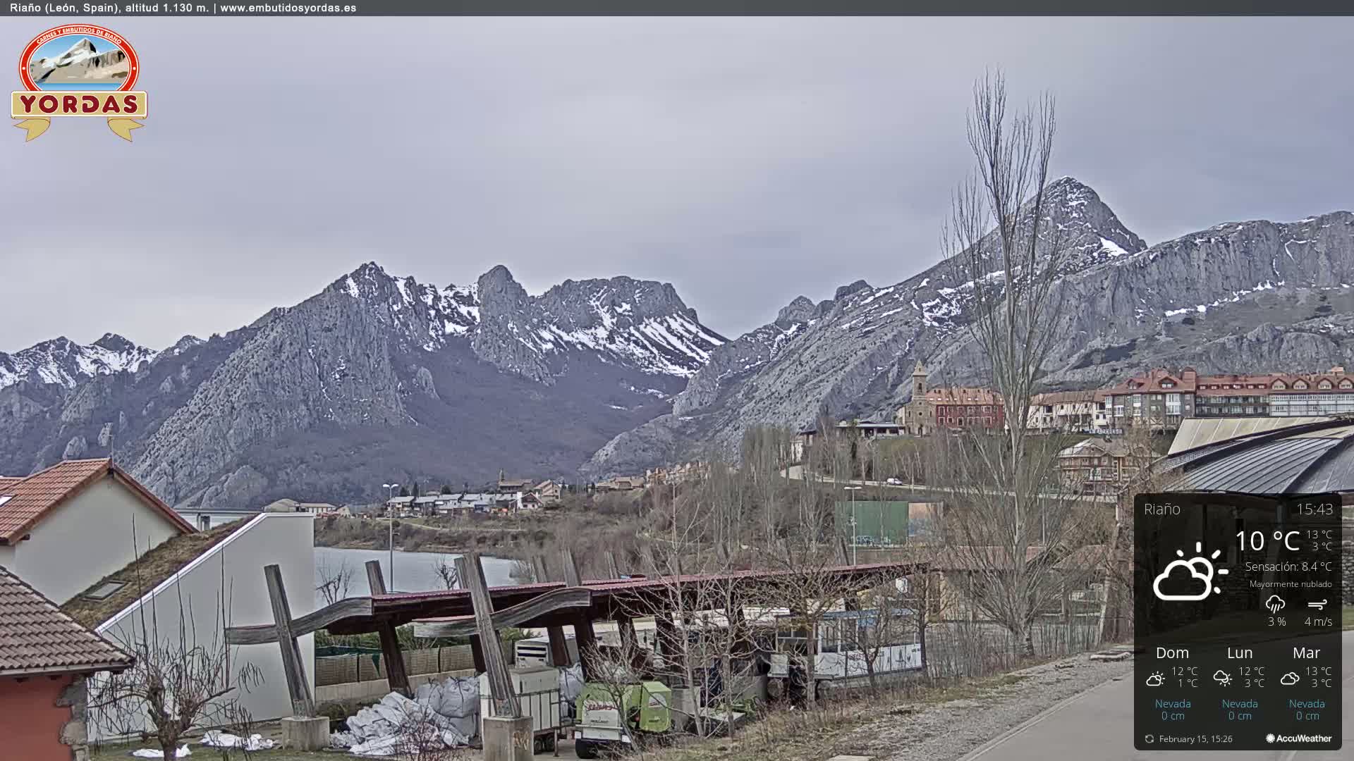 A village nestled in a valley, overlooked by snow-capped mountains under a mostly cloudy sky.