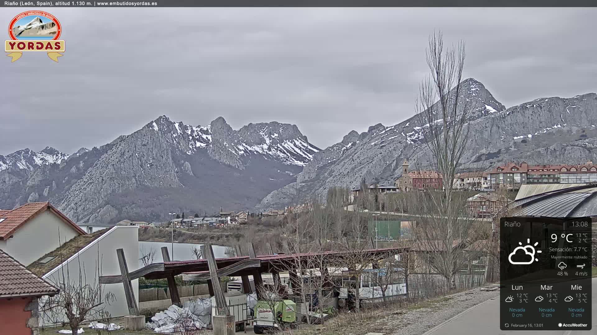 A partly cloudy view of a snow-dusted mountain range overlooking a town with buildings and a covered structure in the foreground.