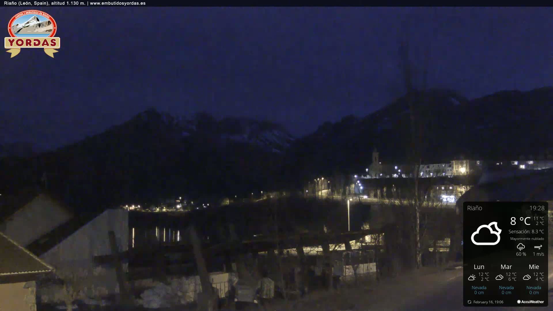 A nighttime view of a small town nestled in a valley between dark mountains under a mostly cloudy sky.