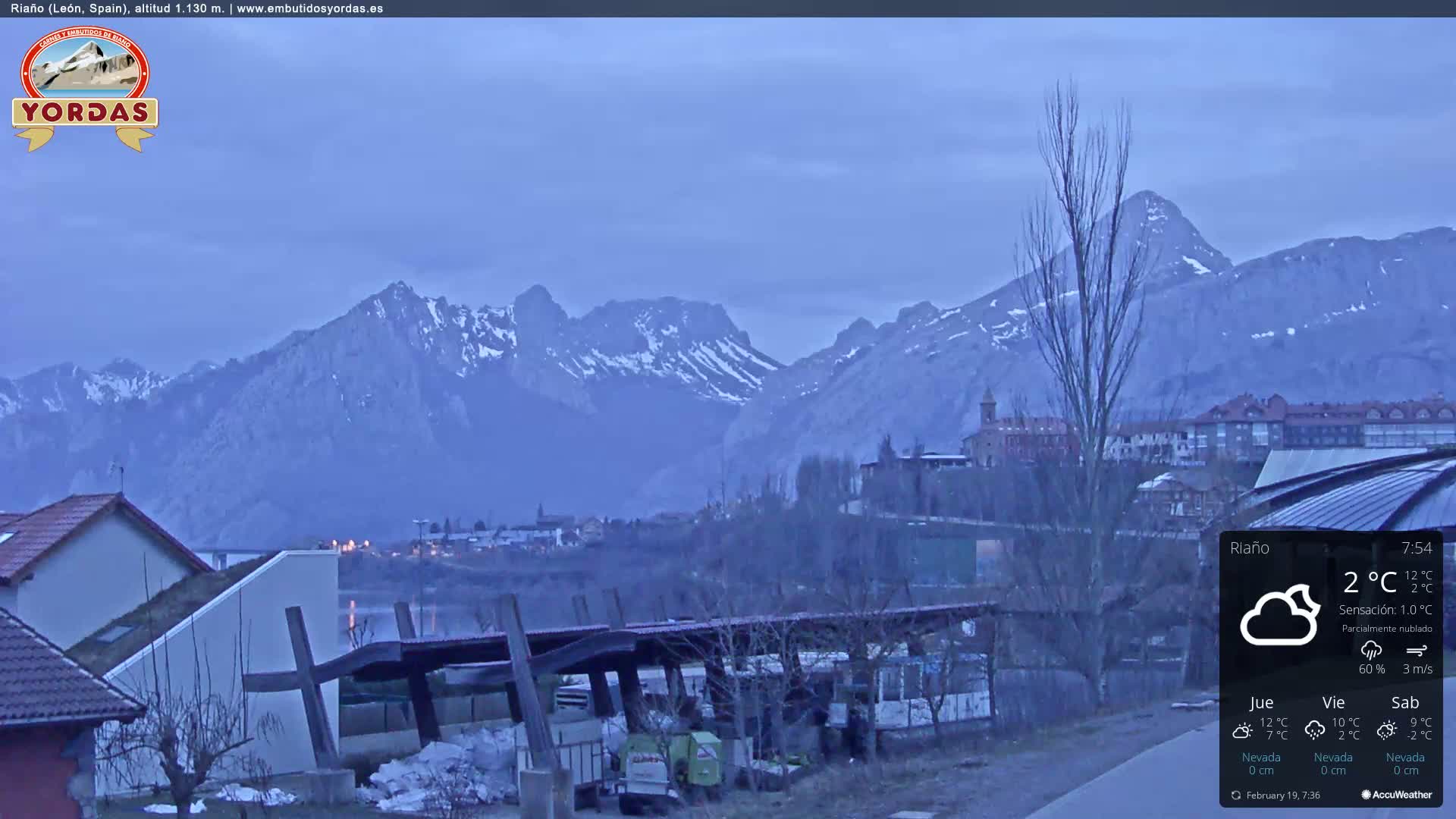 A partially cloudy twilight scene shows snow-capped mountains in the background, overlooking a town and buildings with bare trees in the foreground.